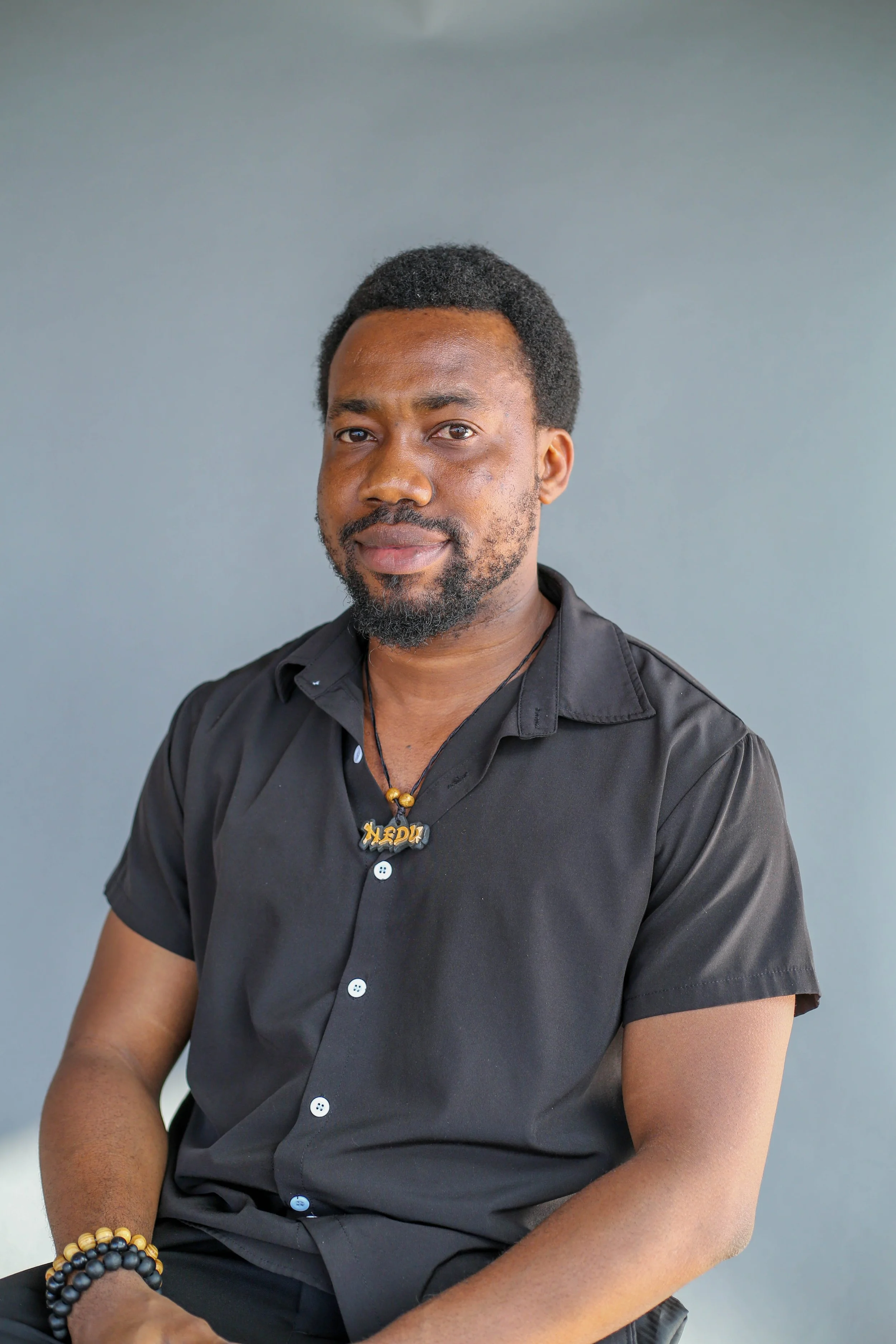 Portrait of a man with a beard and short black hair, wearing a black shirt, sitting against a plain gray background.