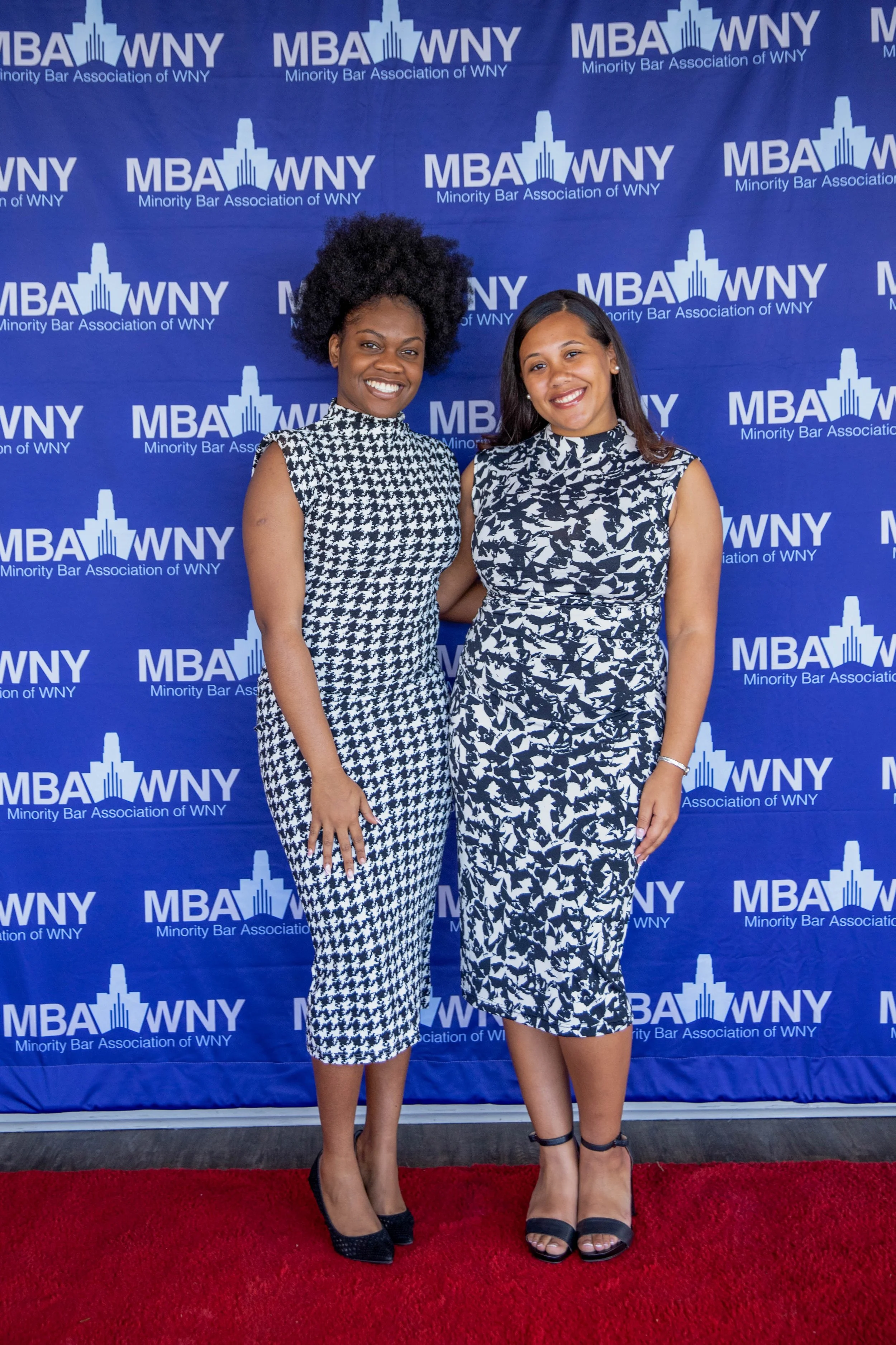 Two women standing in front of a blue backdrop with white text and logos of the Minority Bar Association of WNY, smiling at the camera. They are dressed in black and white patterned dresses, black shoes, and one has black strappy heels while the othe