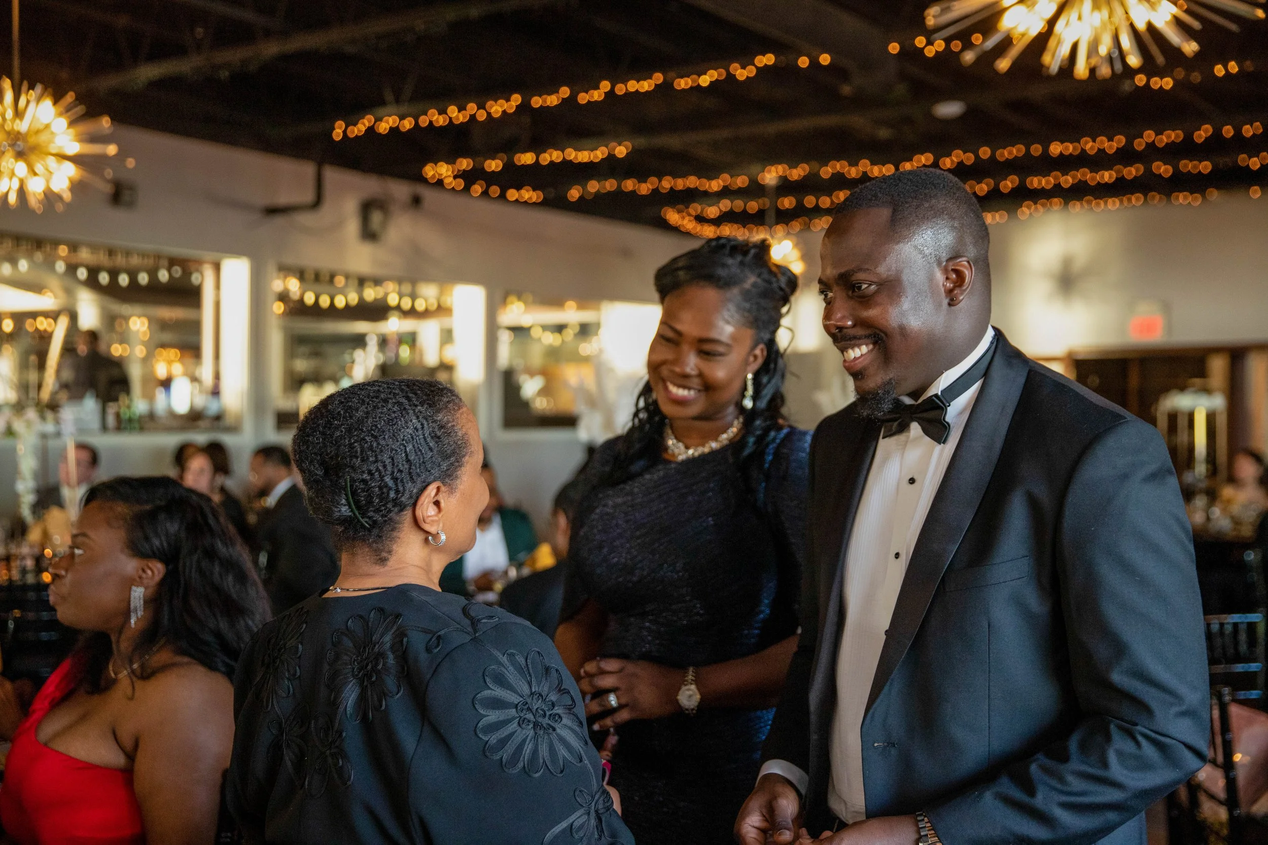 A group of people dressed in formal attire enjoying a social event or celebration in a decorated venue with string lights and festive decorations.