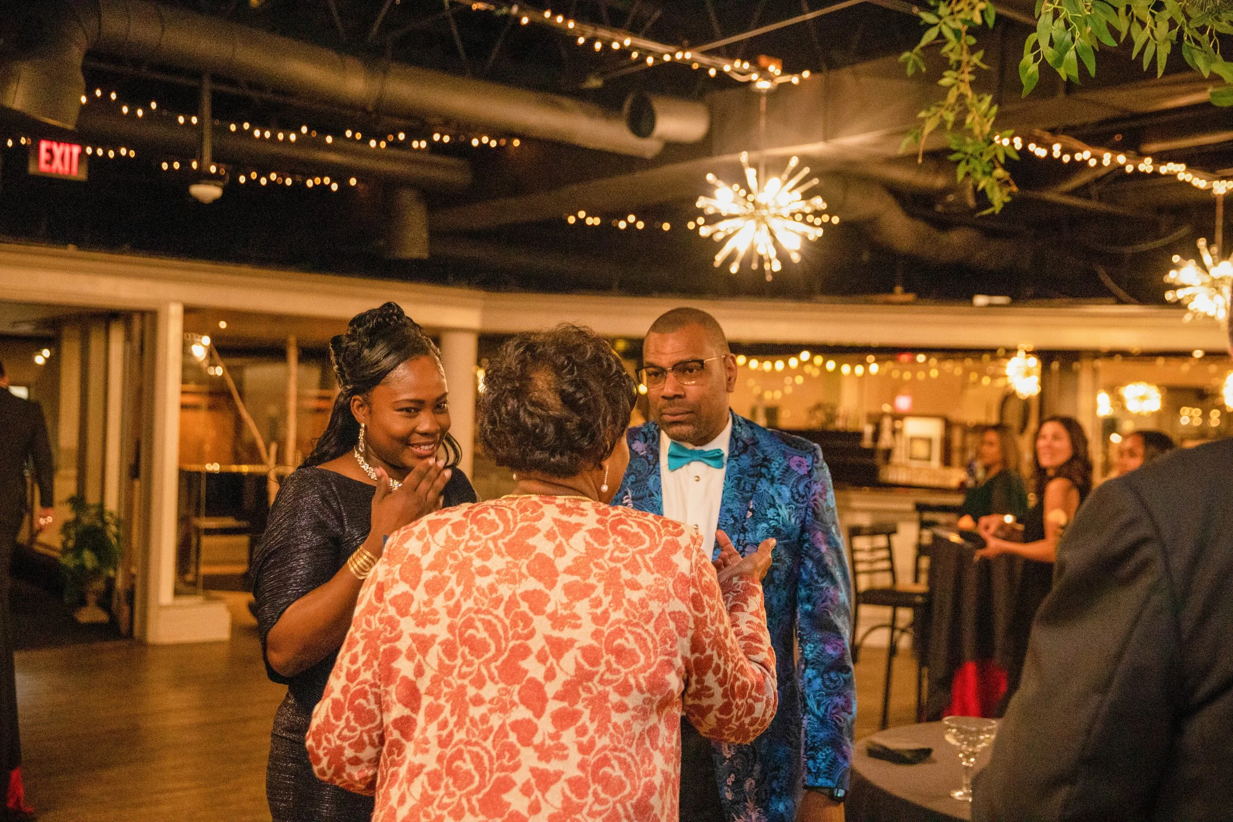 People socializing at a party or gathering in a dimly lit venue with hanging string lights and decorative fixtures.