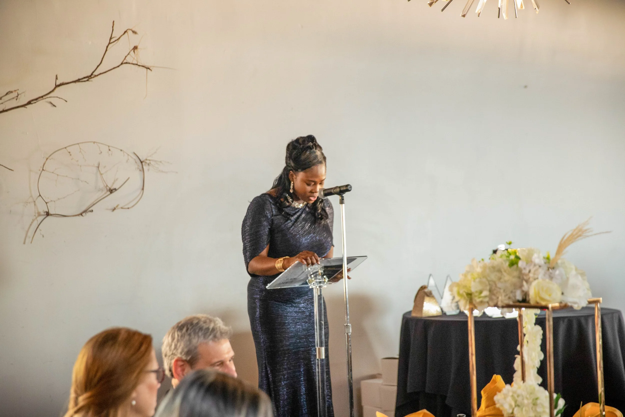 Woman in a dark, sparkly dress giving a speech at a podium, with a microphone, in a decorated indoor venue with floral arrangements and people seated nearby.