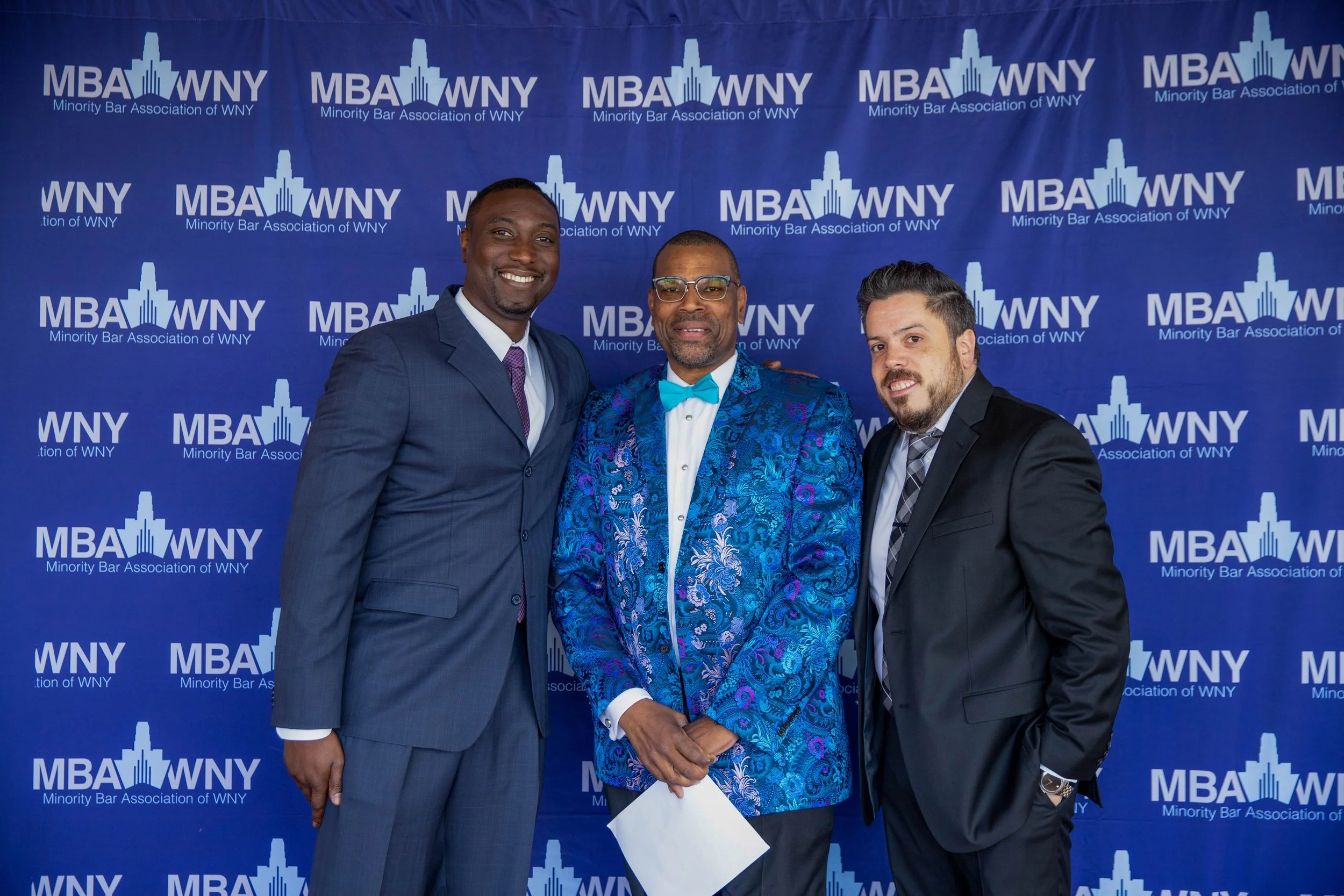 Three men in suits smiling at the camera at an event with a blue backdrop that reads "MBA WNY Minority Bar Association of WNY" and features a city skyline logo.