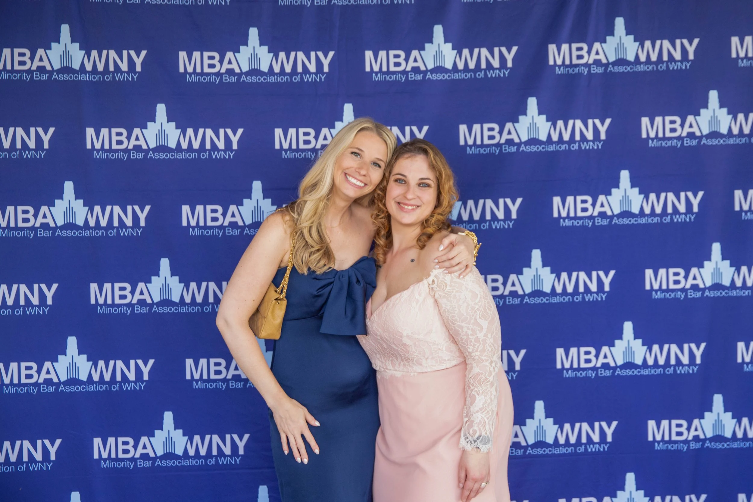 Two women posing in front of a blue backdrop with a repeating logo for the Minority Bar Association of WNY. One woman is wearing a navy blue dress with a gold purse, and the other is wearing a light pink lace dress. Both women are smiling and have th