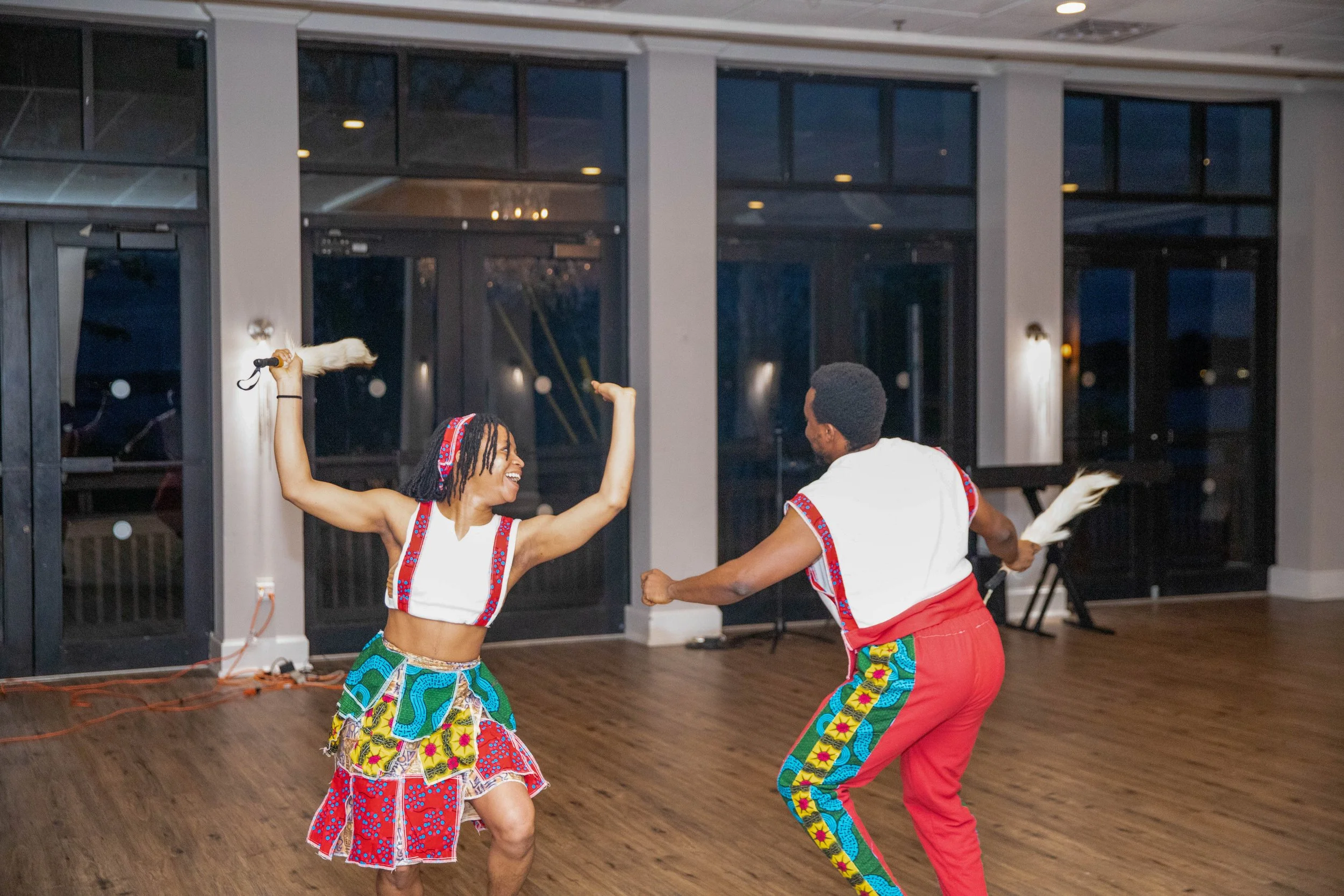 Two people dancing indoors with large windows in the background. They are wearing colorful, patterned clothing and appear to be enjoying a lively dance.