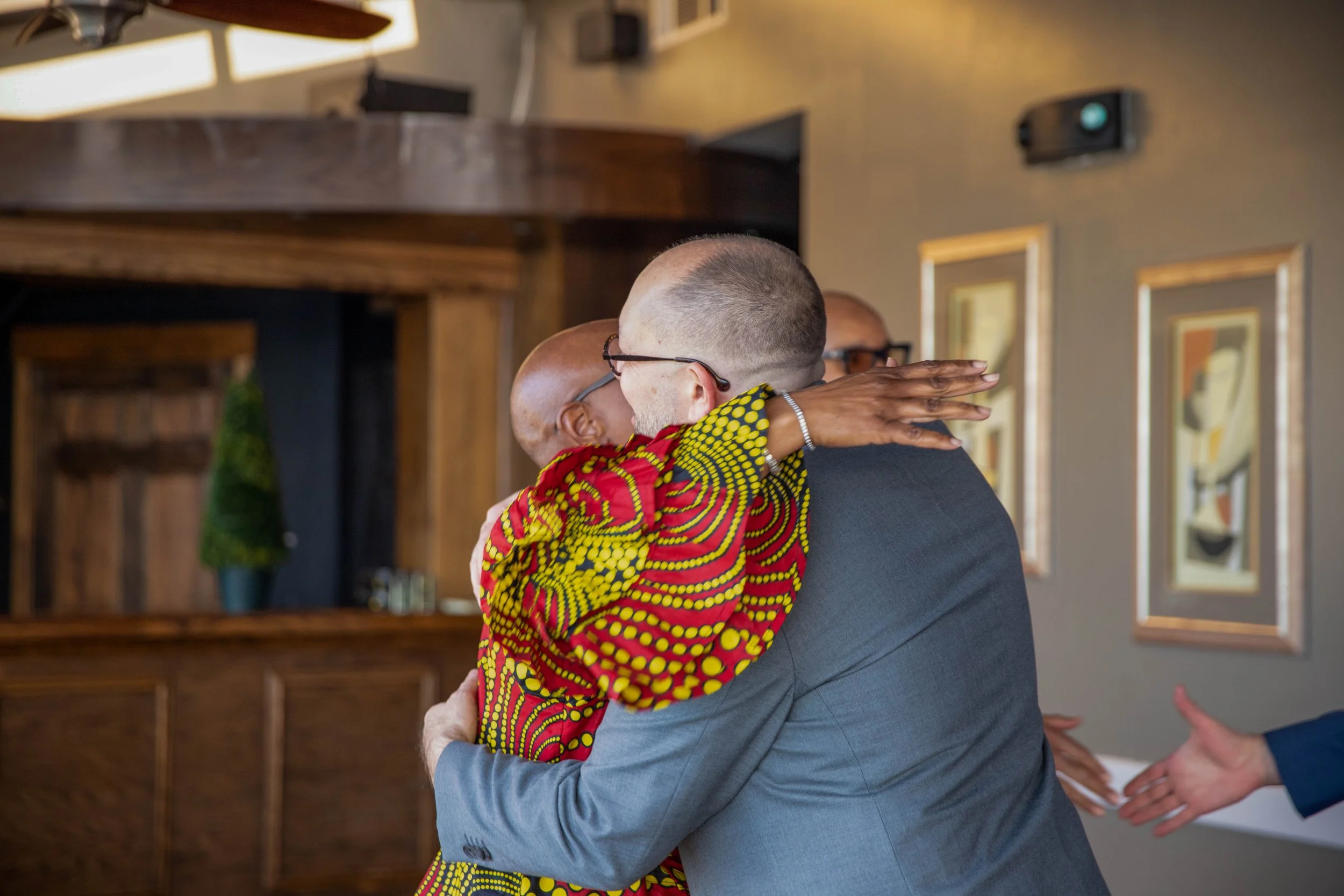 Two people hugging, one wearing glasses and a bold, colorful patterned shirt, the other in a grey suit, in an indoor setting with framed artwork on the wall.