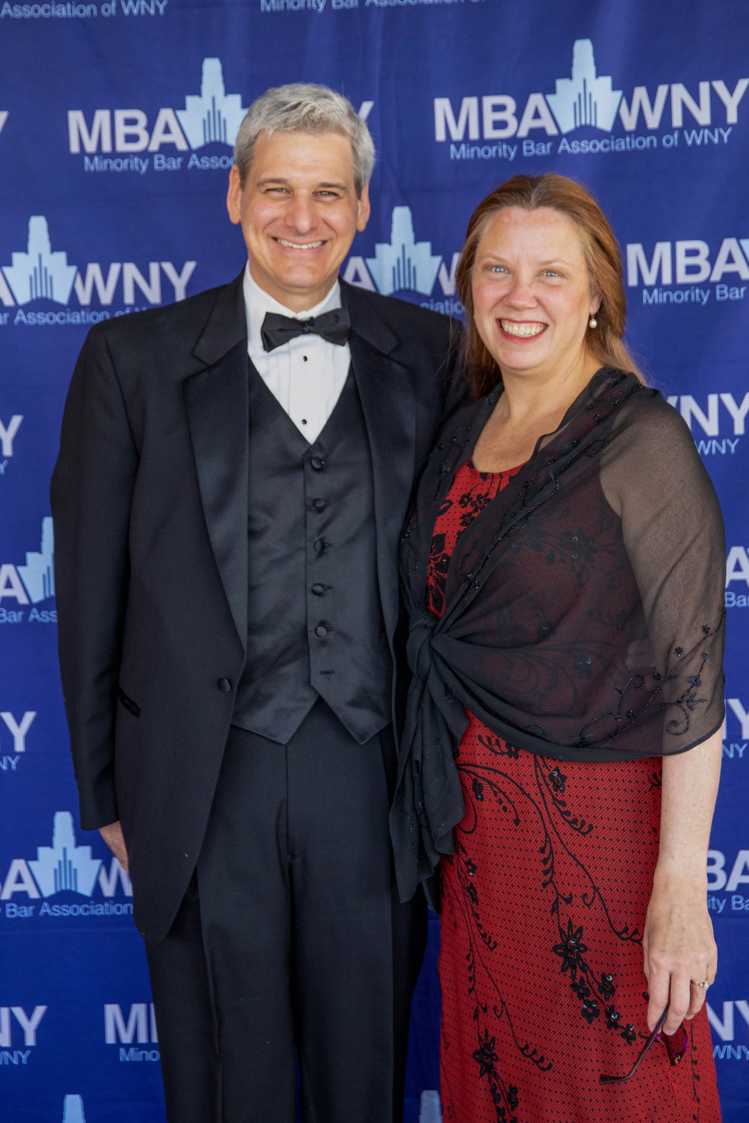 A man and a woman dressed formally, smiling at the camera, standing in front of a blue backdrop with the logo and text for the Minority Bar Association of WNY.