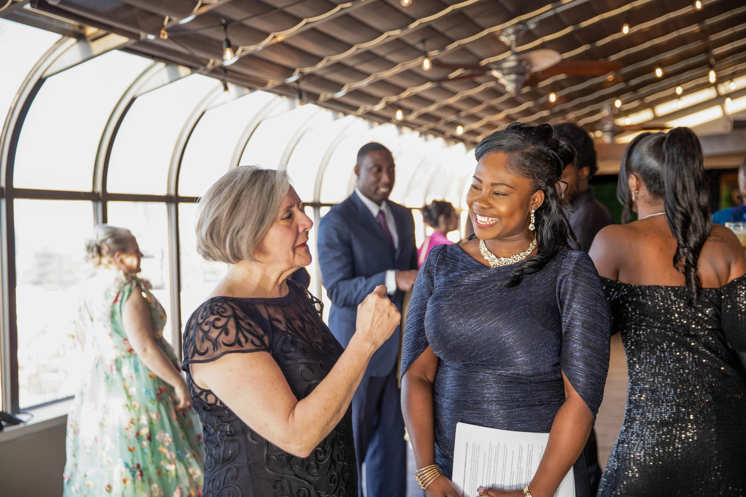 People socializing at a formal indoor event, two women are conversing and smiling, one holding a paper, others mingling in the background, large windows with natural light, string lights hanging from the ceiling.