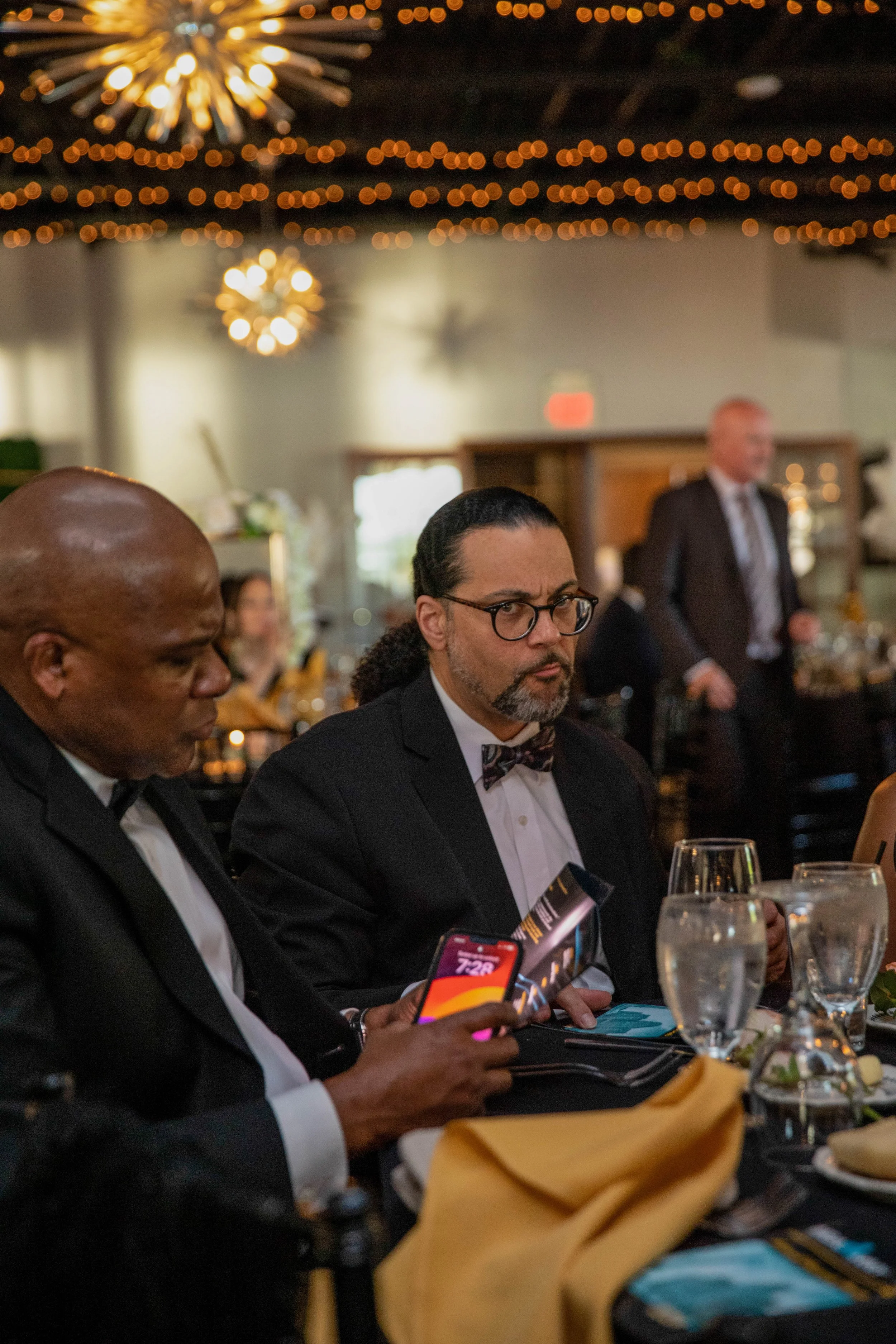 People dressed in formal attire dining at a banquet, with glasses of water, menus, and plates of food on a table, decorated for a celebration or event, with string lights and chandeliers in the background.