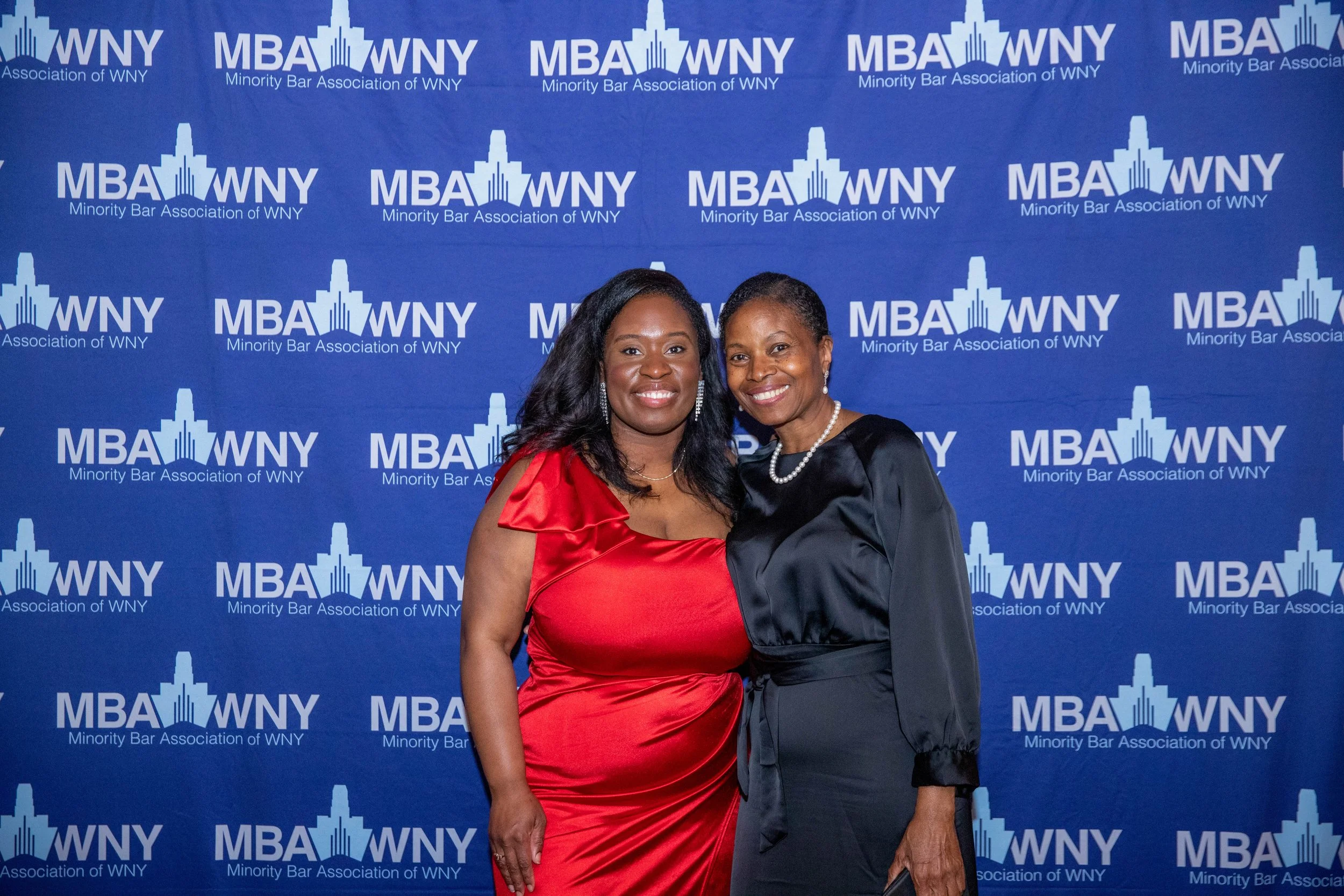 Two women posing in front of a blue backdrop with the logo and text 'MBA WNY Minority Bar Association of WNY'. One woman in a red dress and the other in a black dress with pearls.