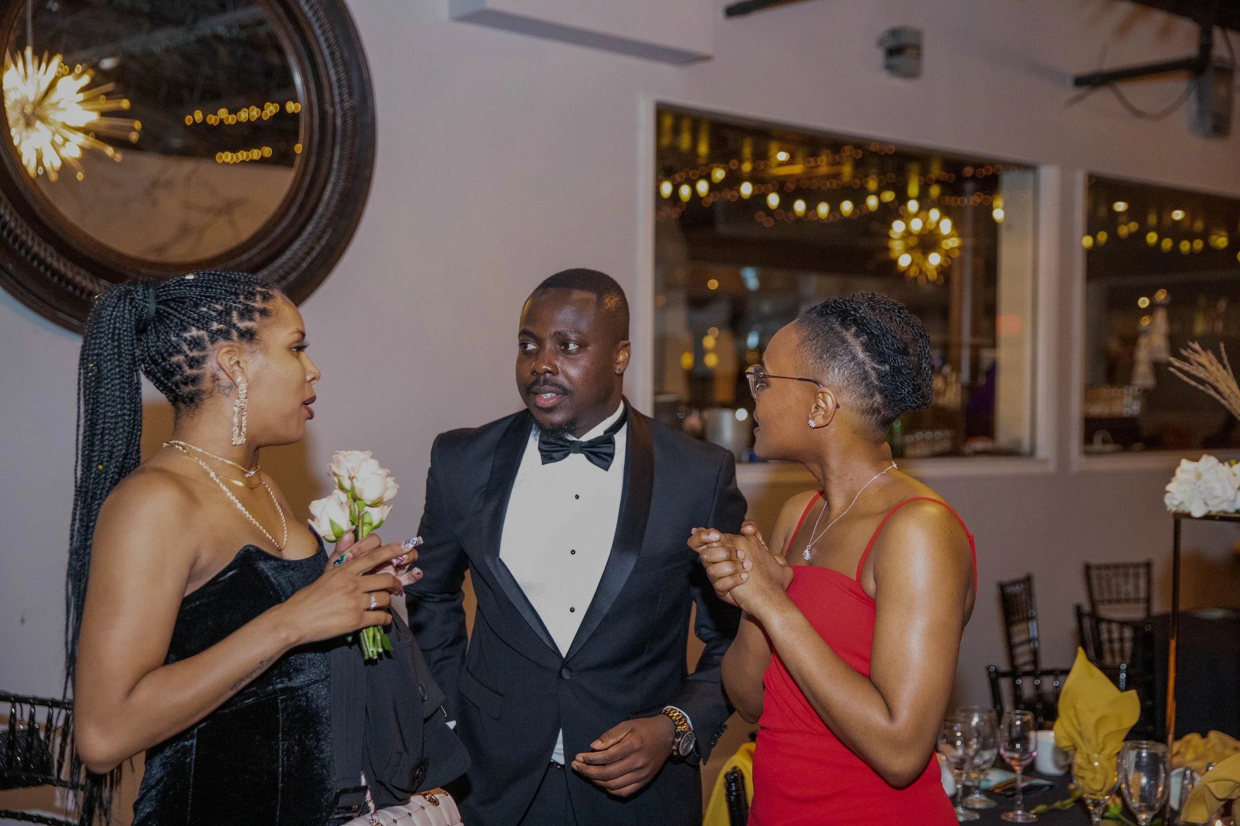 Three people are having a conversation at a formal event, with two women and one man in a tuxedo. The woman on the left is wearing a black dress, the man is in the middle, and the woman on the right is in a red dress. They appear engaged and attentiv