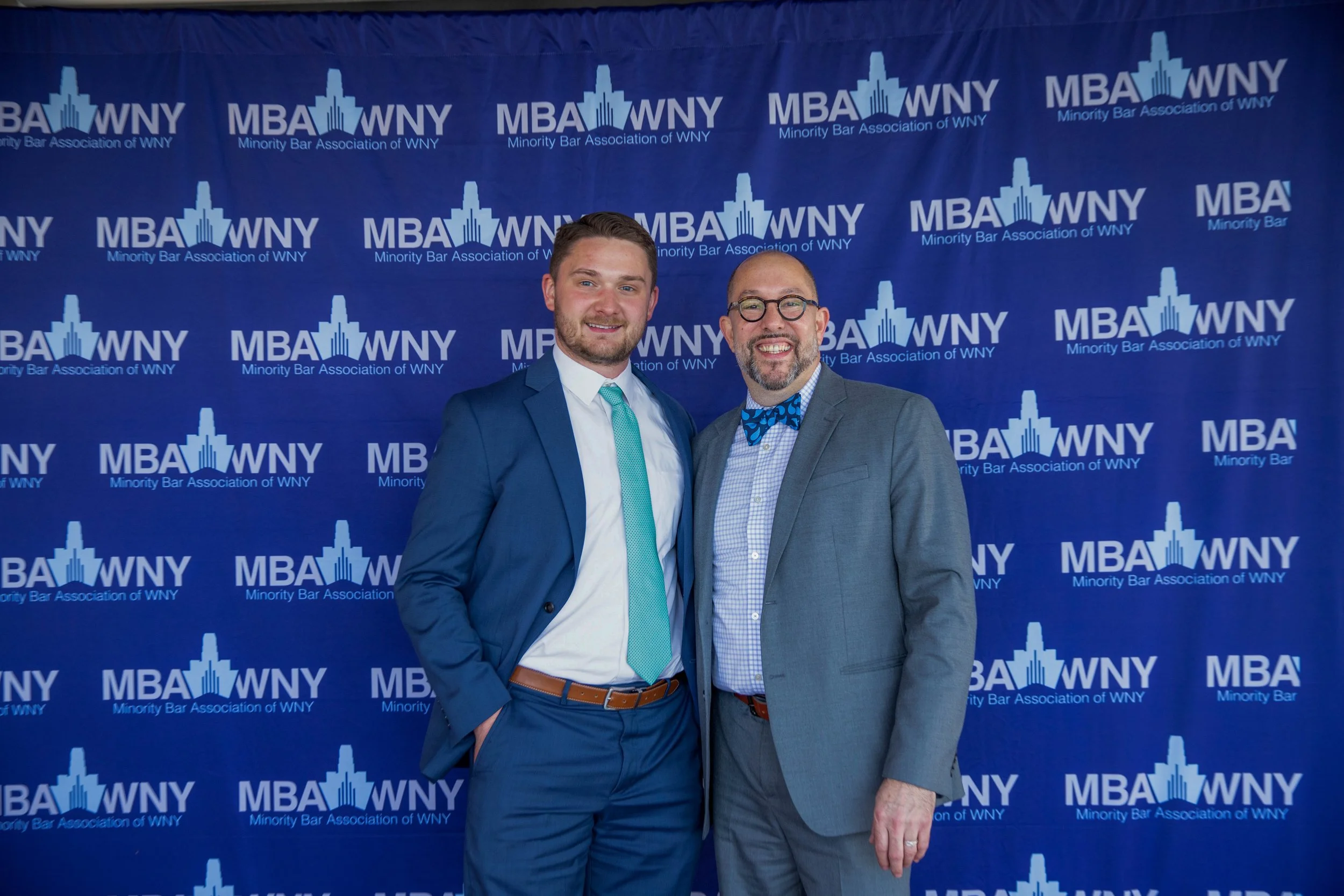 Two men in suits posing in front of a blue backdrop with the logos of the Minority Bar Association of WNY, wearing smiles, one with a teal tie and the other with a blue patterned bow tie.