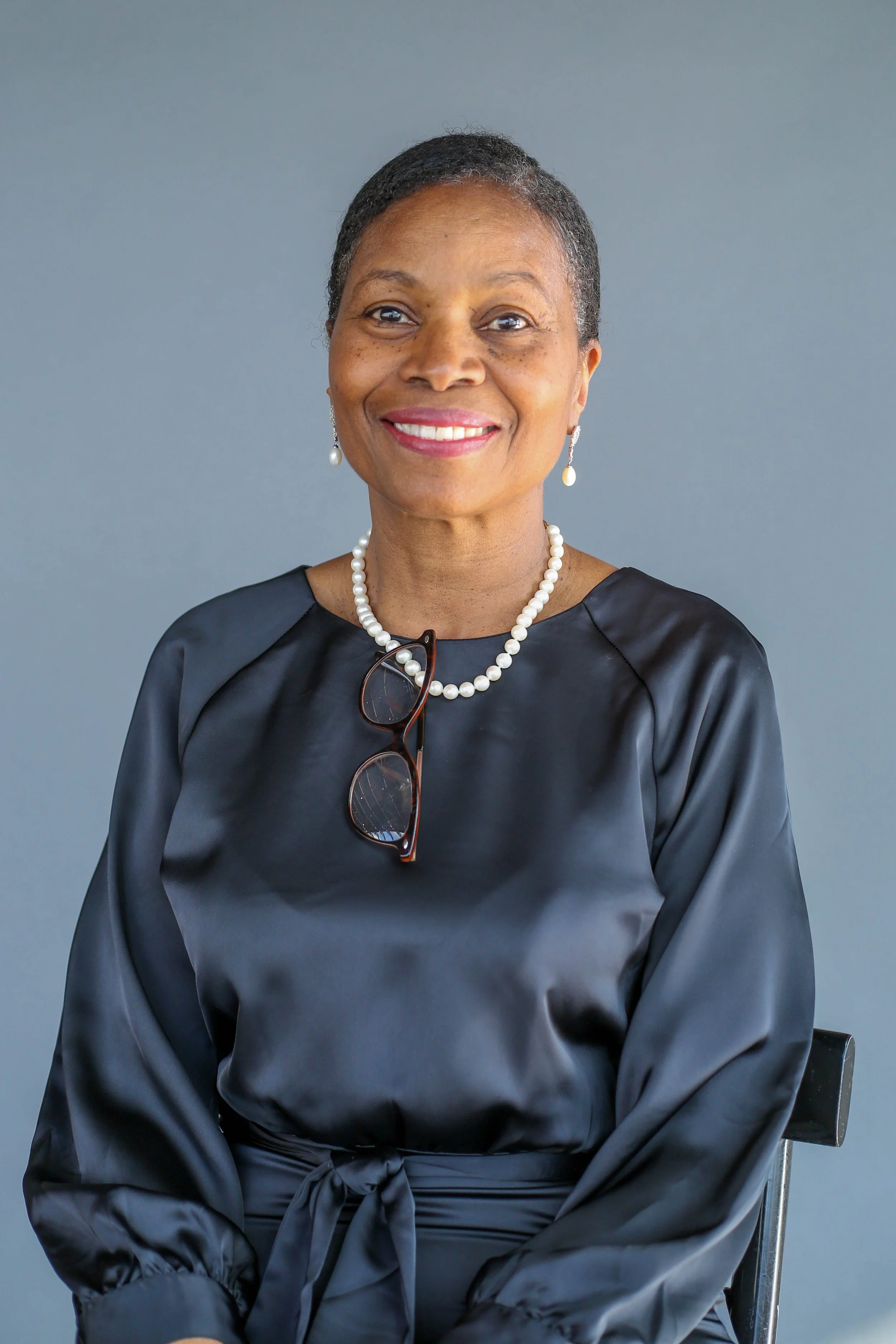 A woman with dark short hair, wearing pearl earrings and a pearl necklace, smiles at the camera. She is dressed in a shiny, dark gray satin blouse with a knot at the waist and has glasses hanging from her neck. She is sitting on a black chair against