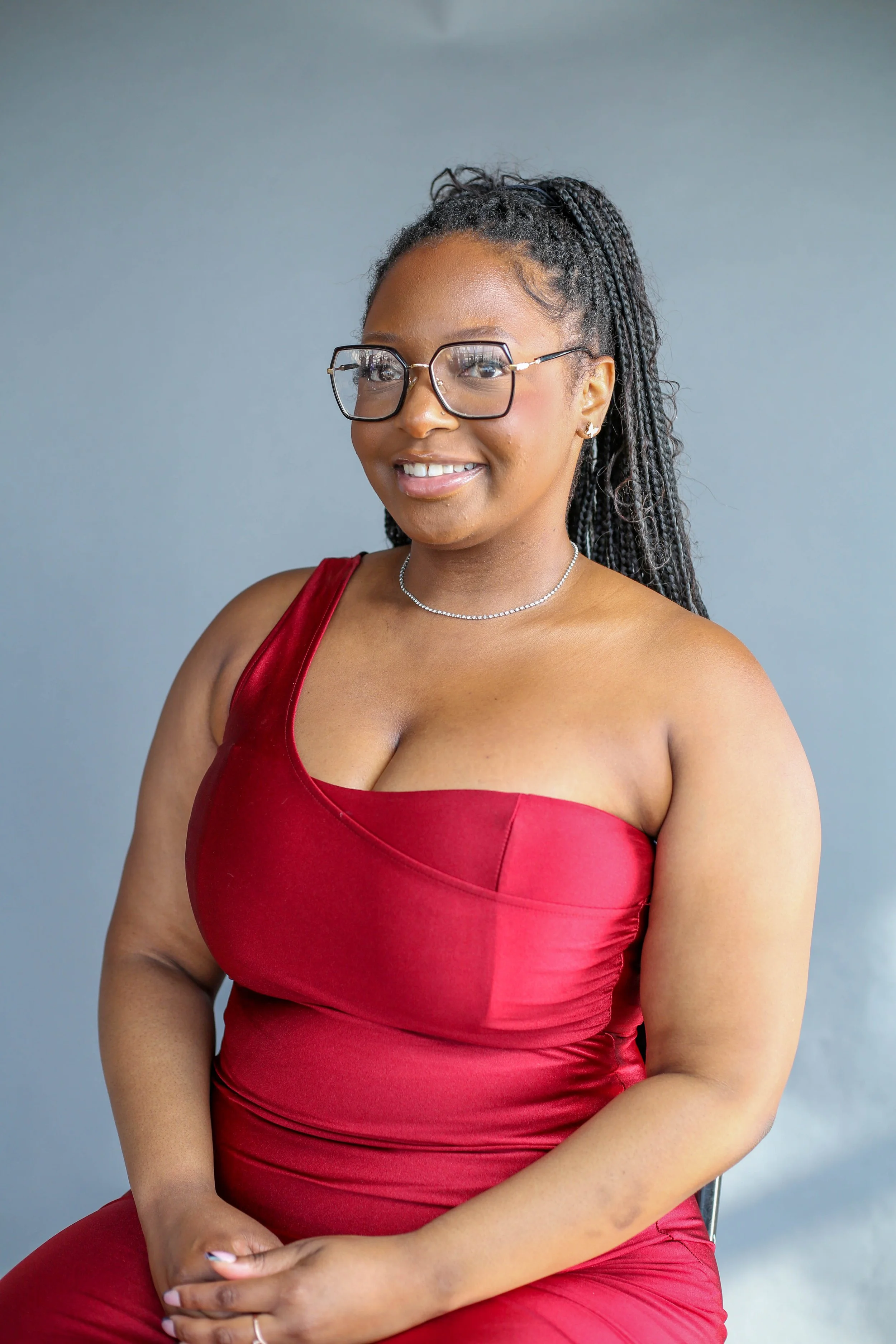 A woman with dreadlocks, wearing glasses, a red satin dress, jewelry, smiling, sitting against a blue-grey background.