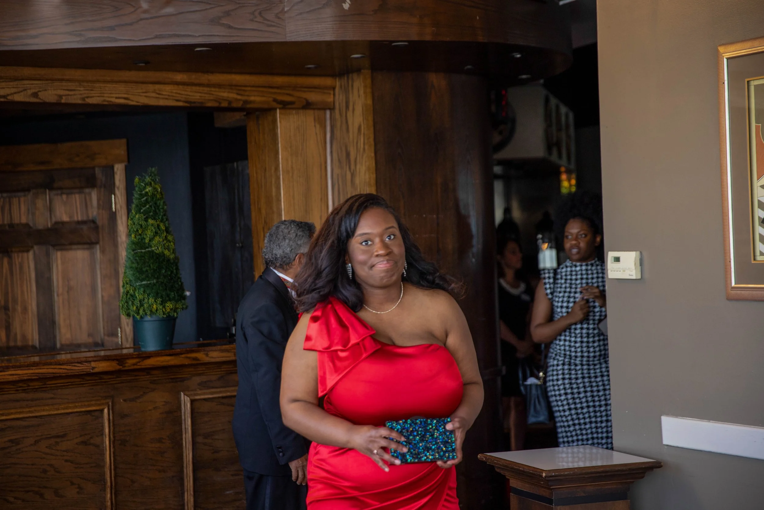 A woman in a red dress holding a beaded clutch, standing indoors at what appears to be a formal event, with other people and wooden decor in the background.