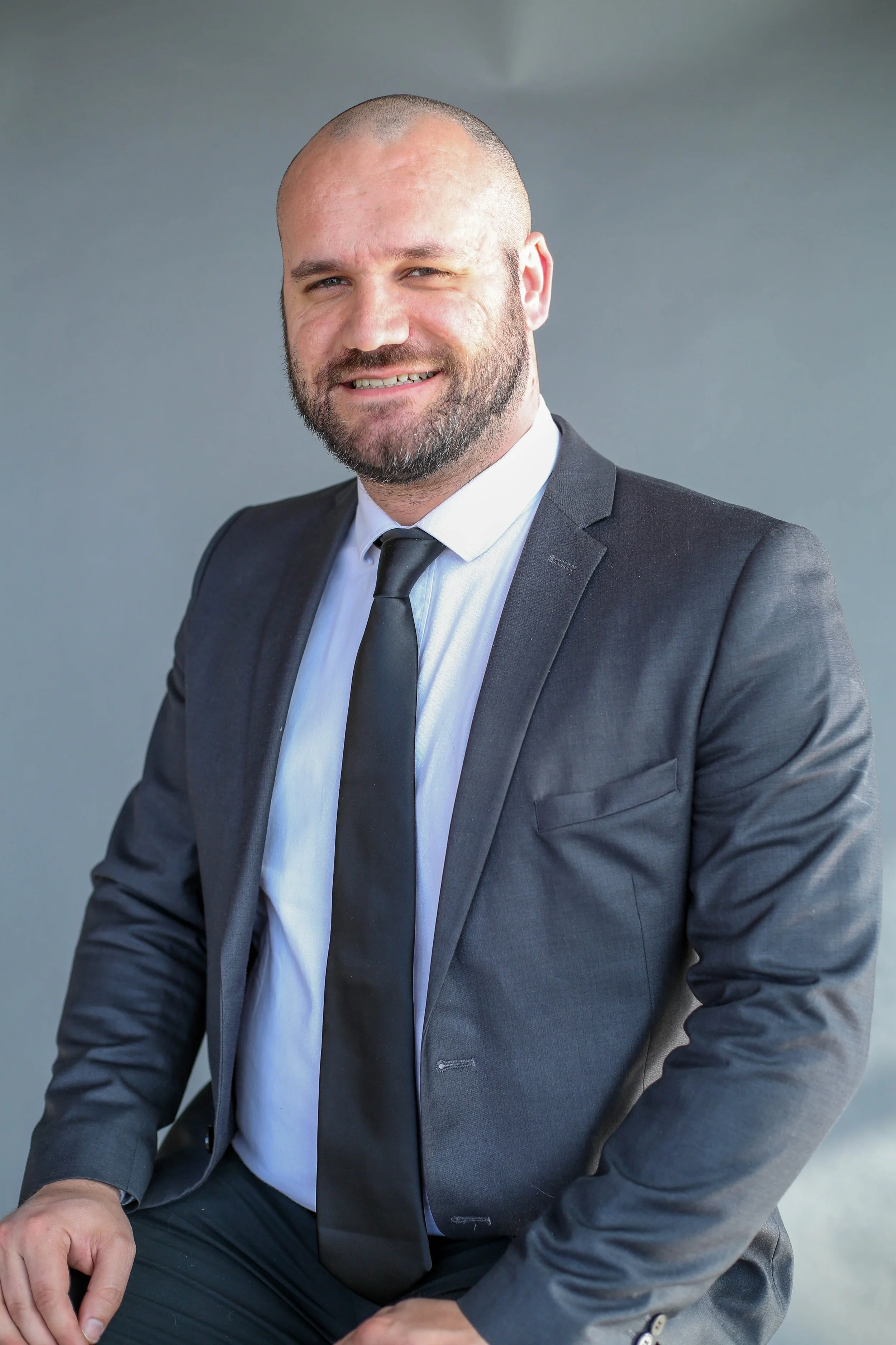 A man in a suit and tie with a beard and shaved head, smiling, seated against a gray background.