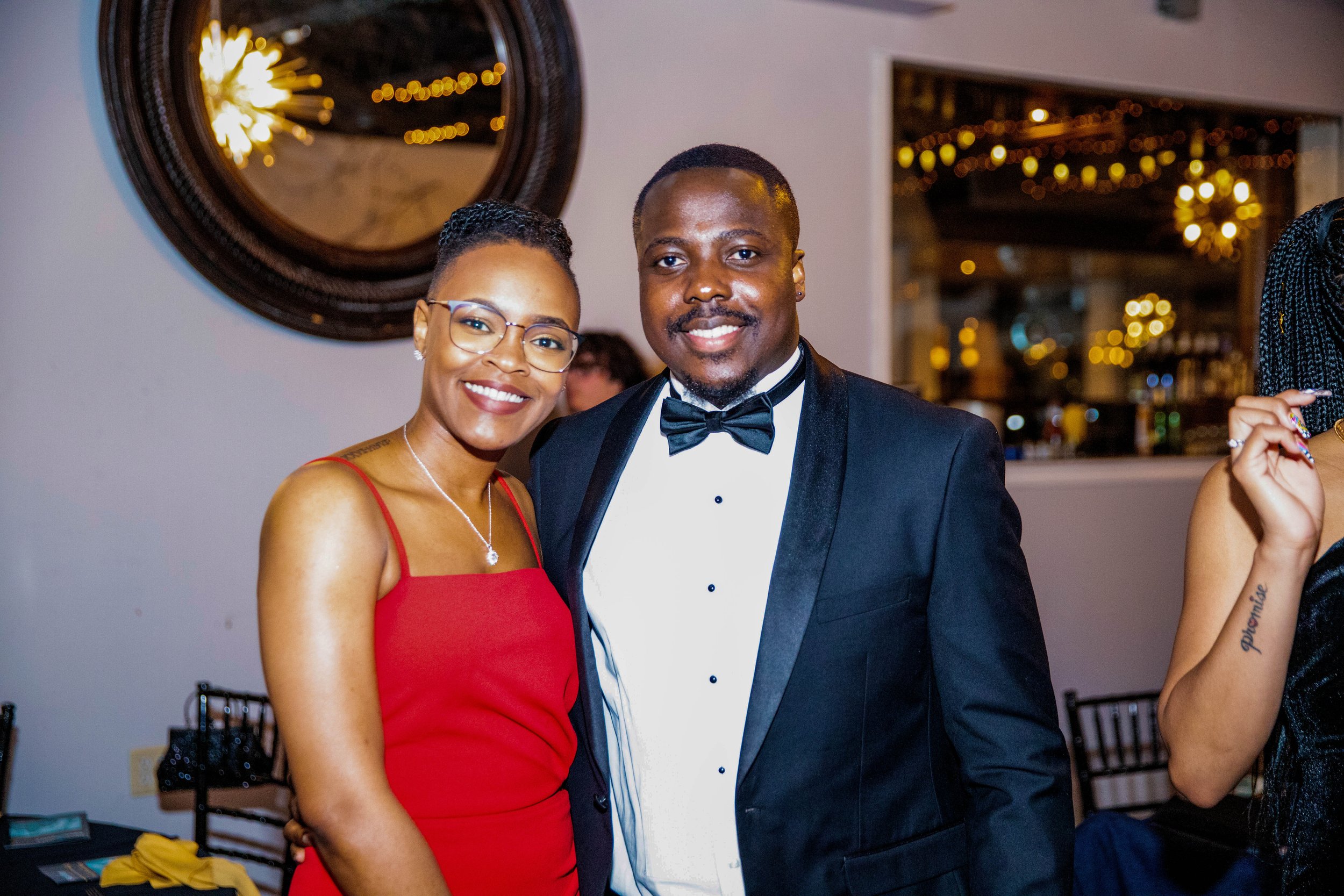 A smiling couple dressed in formal attire at a party or celebration, with the woman wearing a red dress and glasses, and the man wearing a tuxedo. The background features warm lighting and decorative string lights.