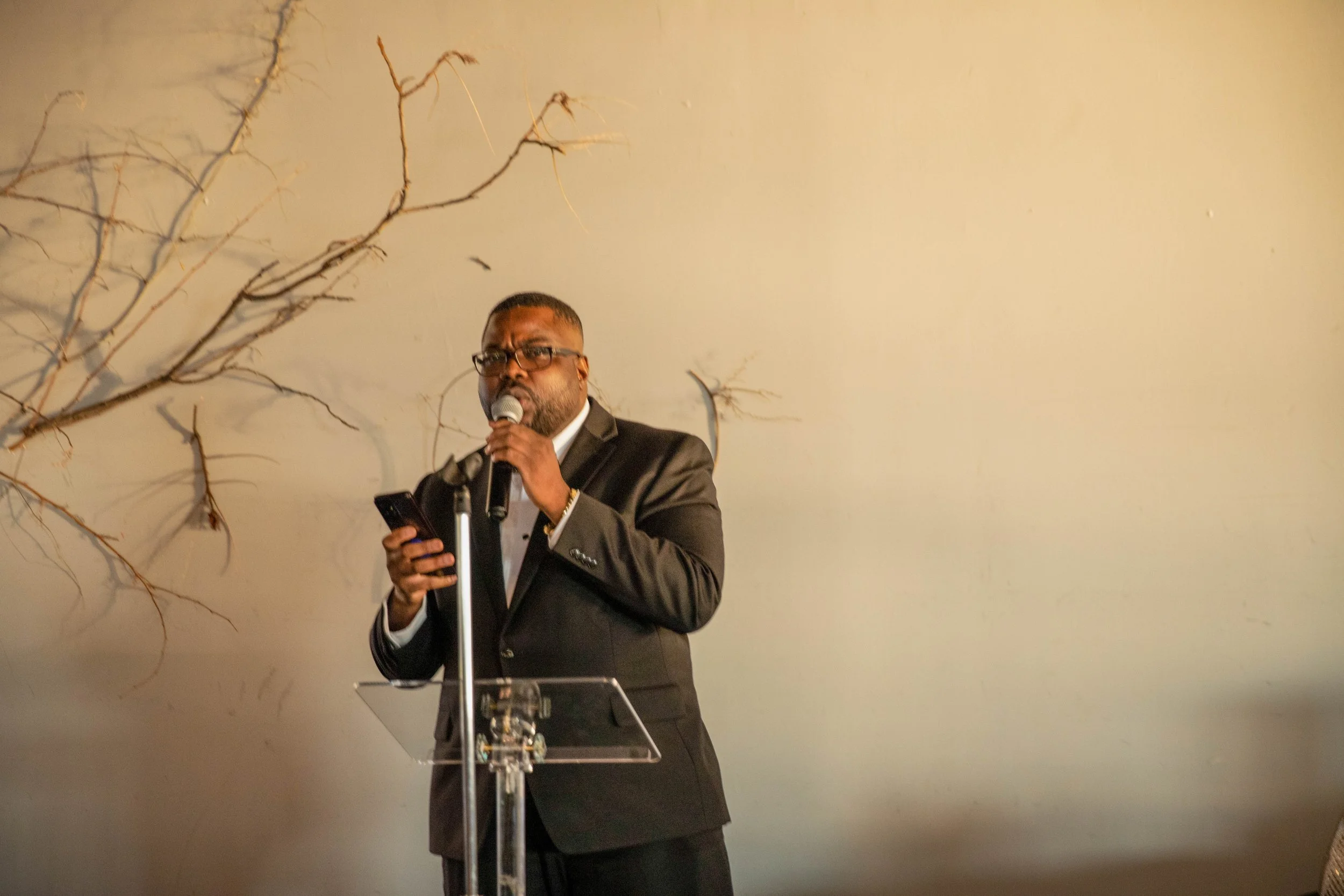 A man wearing a black suit, glasses, and a white shirt is speaking into a microphone at a podium. There is a leafless tree branch to the left of him and a plain, light-colored wall in the background.