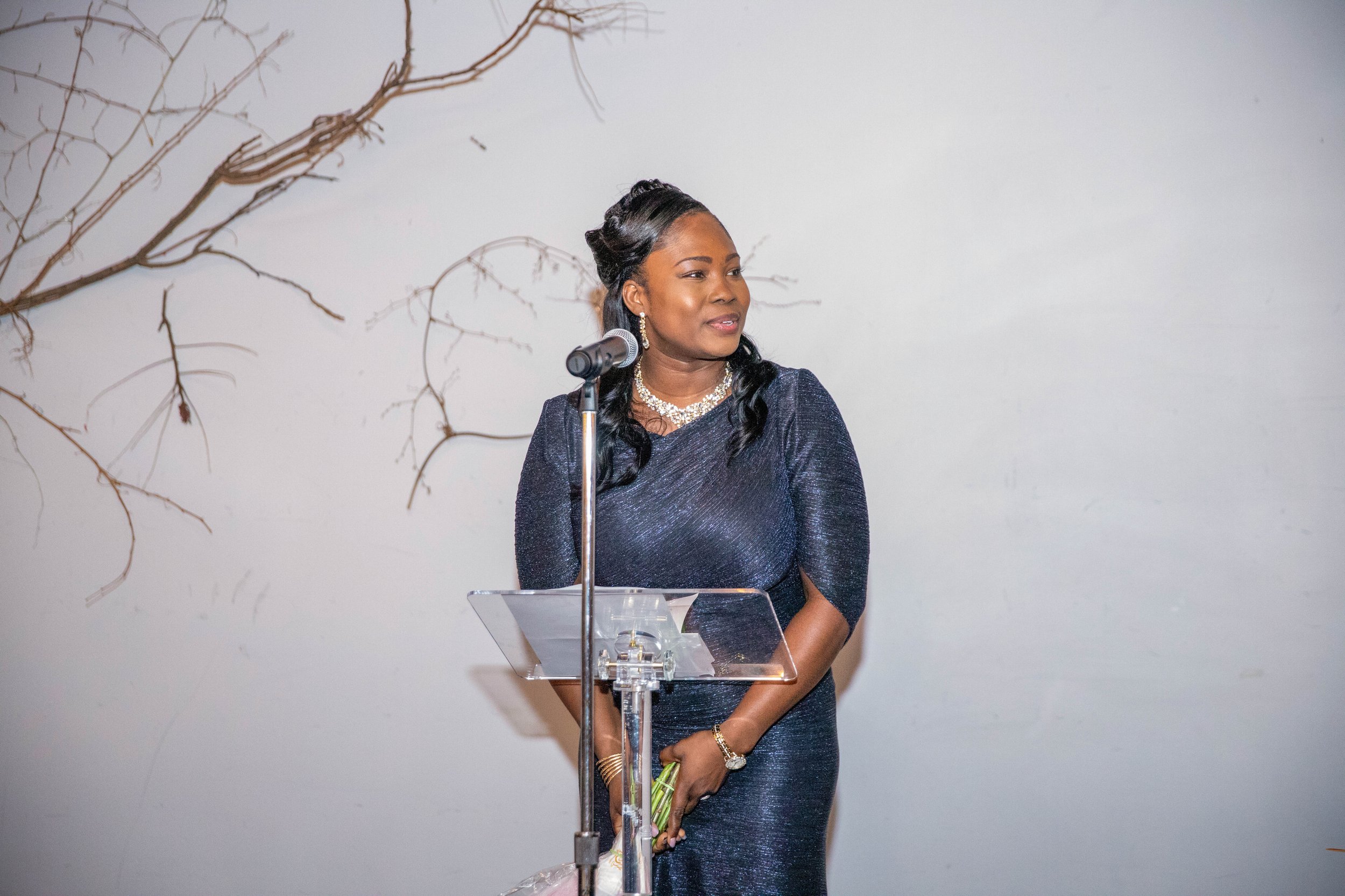 A woman in a sparkly dark blue dress speaking at a podium with a microphone, holding a bouquet of flowers, against a white wall with a decorative branch.