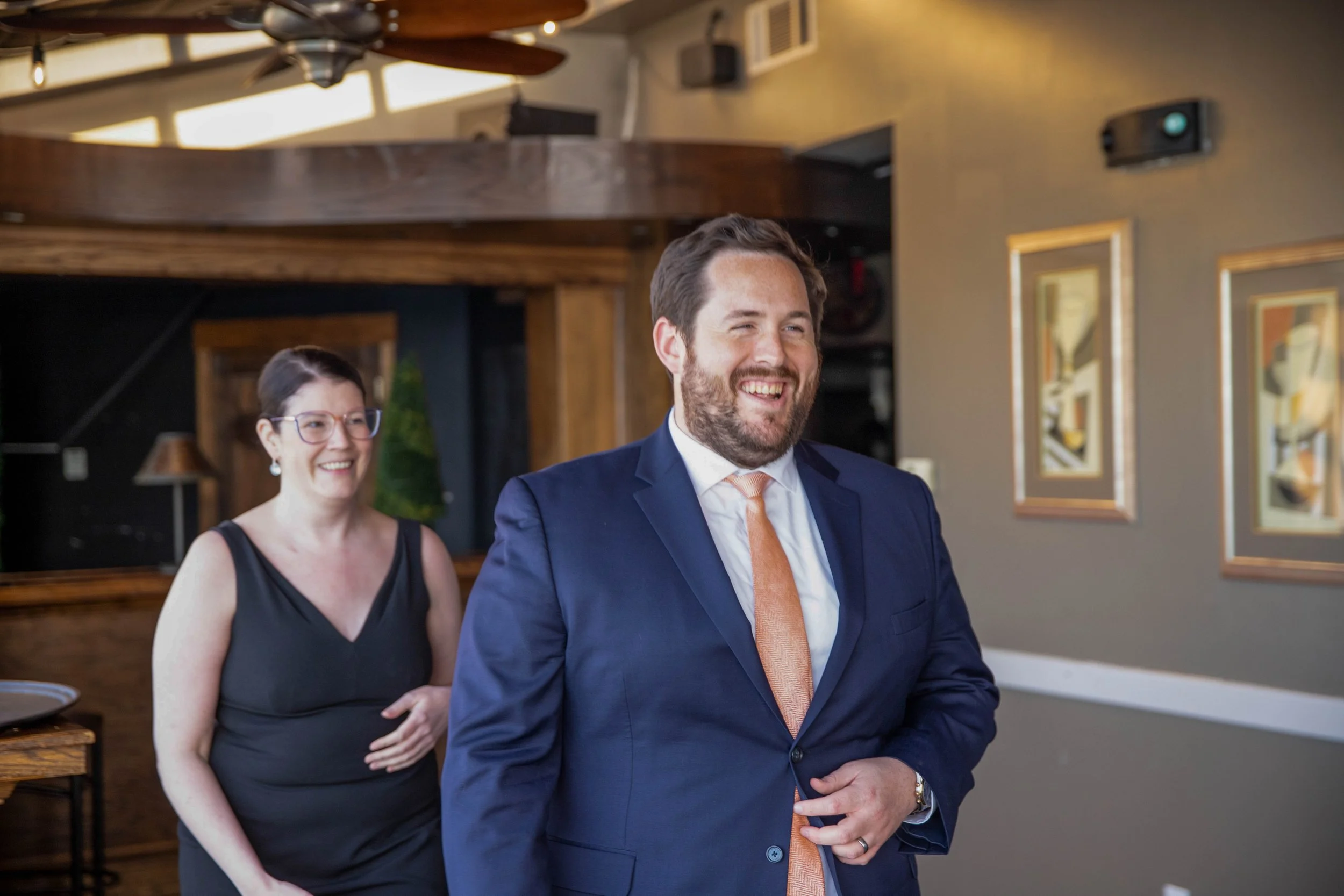 A smiling man in a blue suit with a peach tie standing indoors, with a woman in a black dress and glasses behind him, both appearing happy.