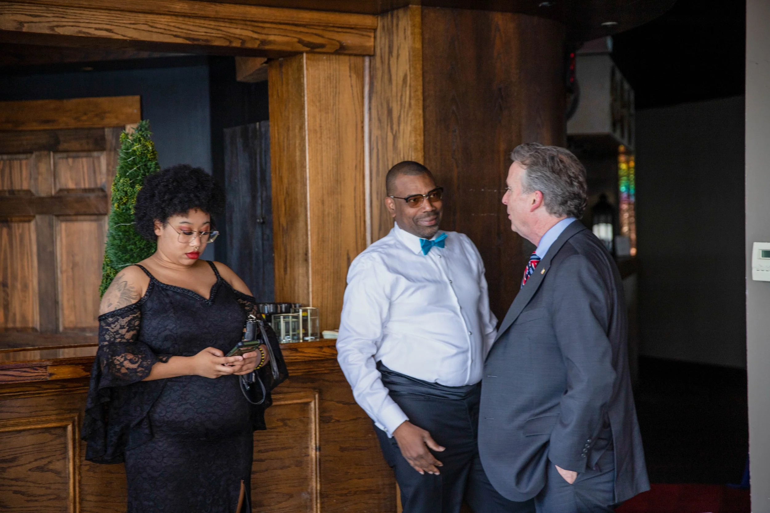 Three people chatting indoors: a woman with glasses and black lace dress looking at her phone, a man in a white shirt and bow tie smiling at a man in a dark suit and tie.