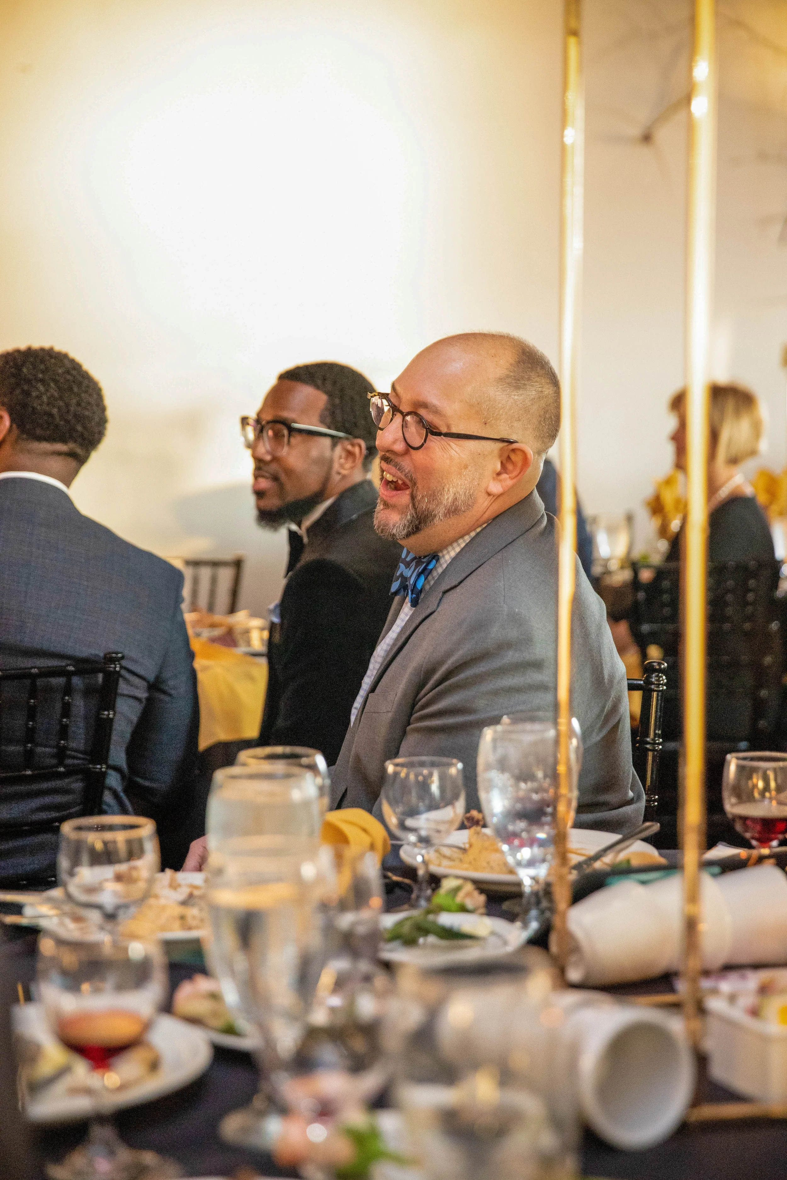 People sitting at a dining table during a social event, with food and drinks on the table, and a brightly lit interior with gold accents.
