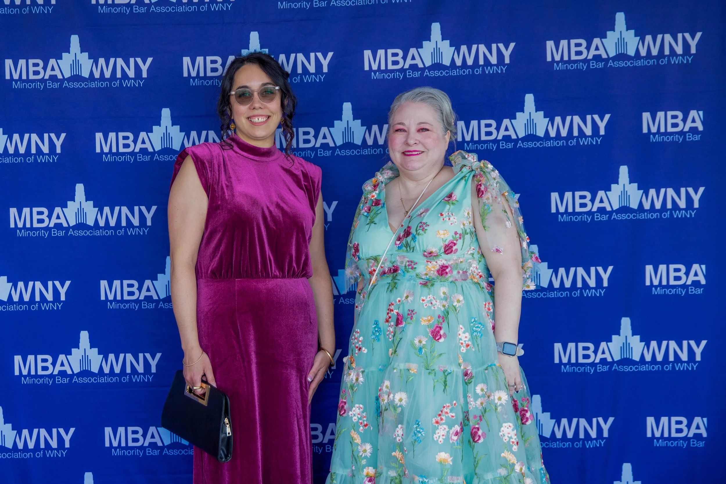 Two women standing in front of a blue backdrop with the logo and text for the Minority Bar Association of WNY. The woman on the left is wearing a purple velvet dress and sunglasses, holding a black purse. The woman on the right is wearing a floral dr