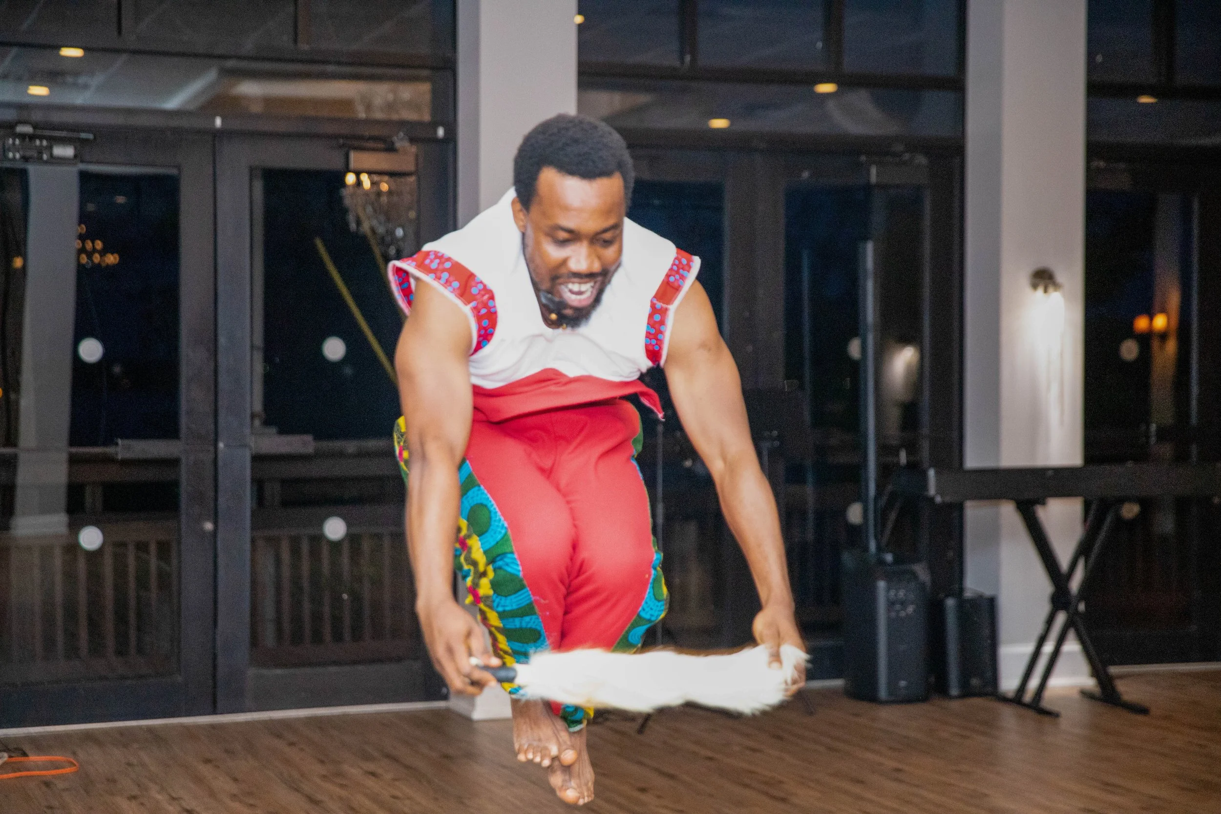 A man with curly hair is dancing or celebrating indoors, wearing colorful clothing with indigenous or traditional patterns, in front of a modern black glass door or window.