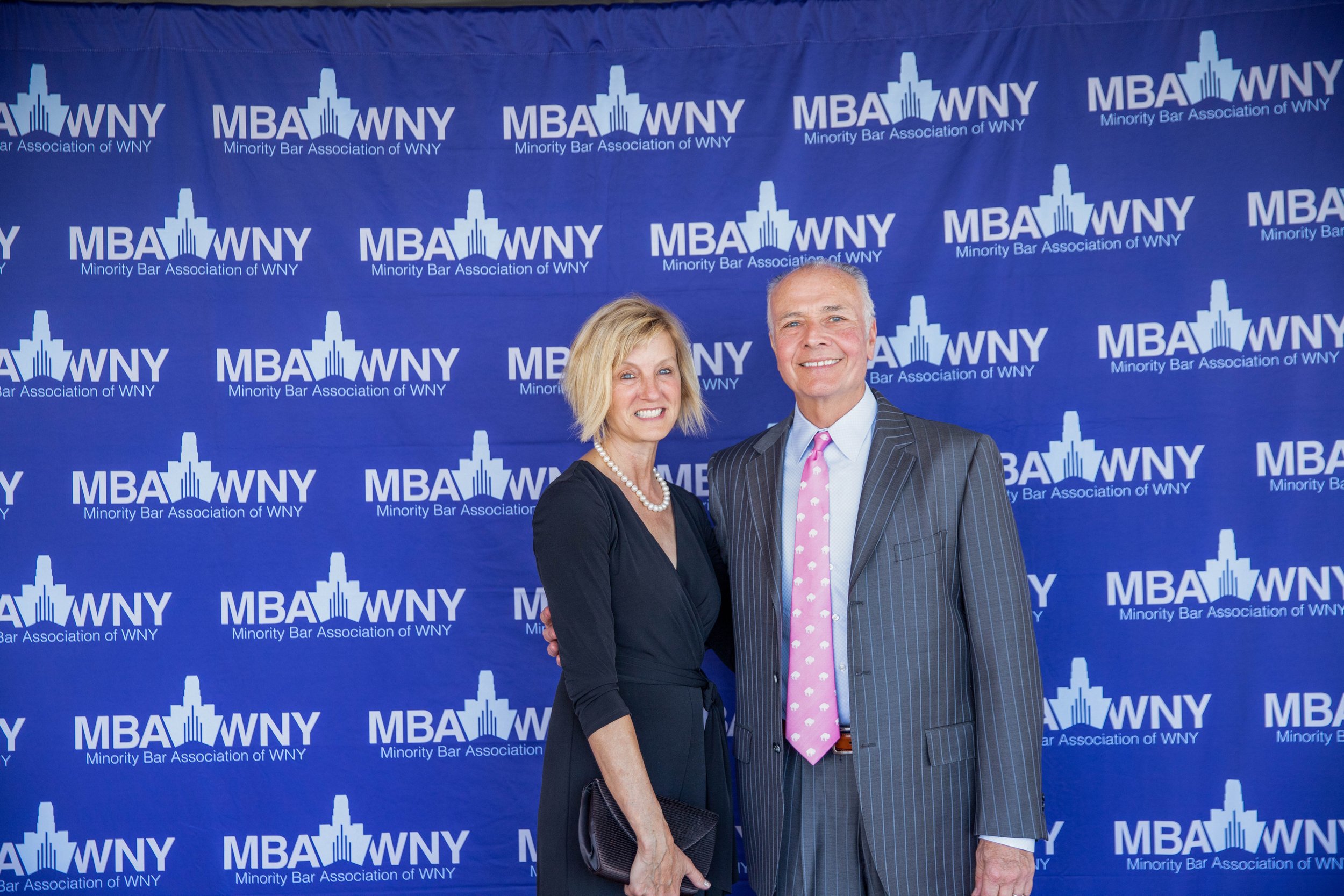 Two people, a woman and a man, standing in front of a backdrop with repeated logos of the Minority Bar Association of WNY. The woman has shoulder-length blond hair and is wearing a dark dress with a pearl necklace. The man has gray hair and is wearin