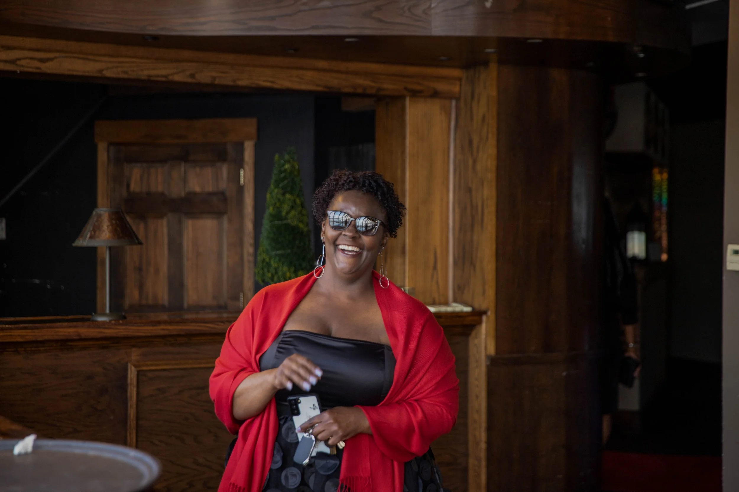 A woman smiling and wearing sunglasses, a red jacket, and earrings, standing in a warmly lit wooden interior space with dark panels and decorative elements.