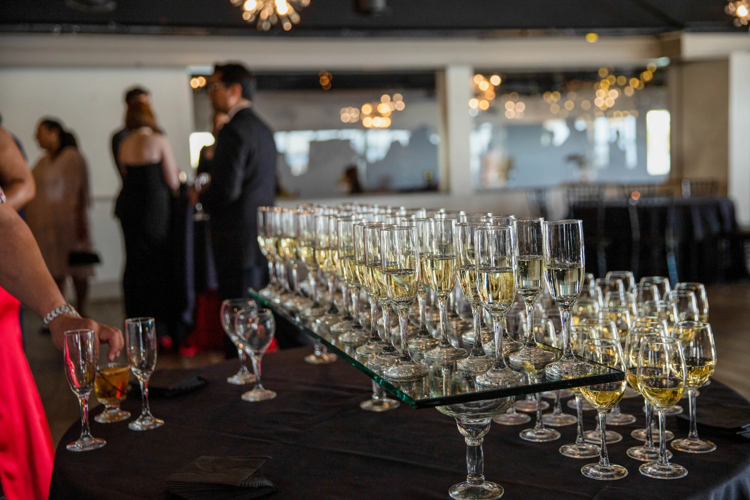 A banquet table with numerous glasses filled with white wine, set up for an event in a decorated indoor venue with hanging string lights and people in formal attire.
