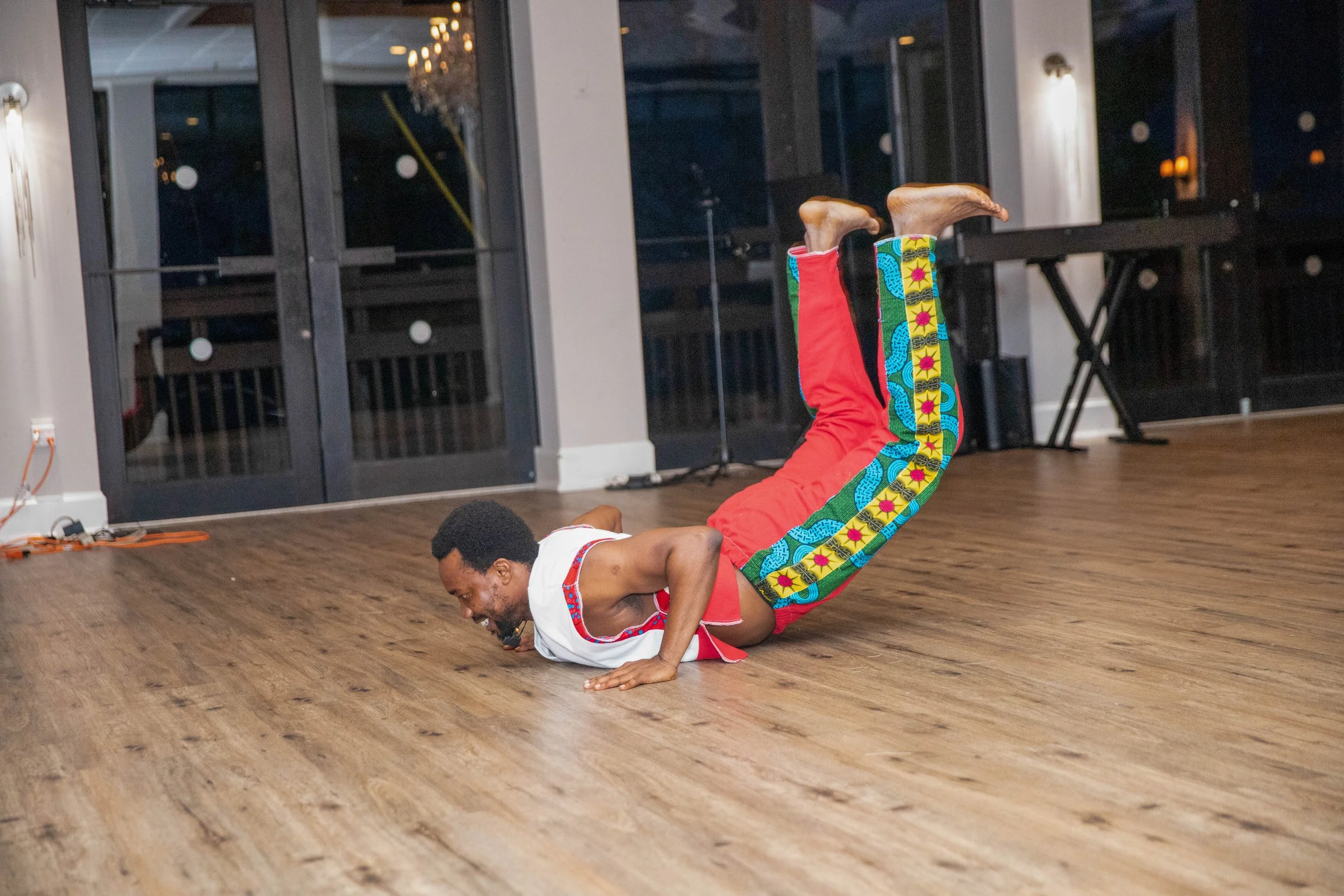 A man wearing colorful pants and a white tank top is performing a yoga pose on a wooden floor indoors, with large windows and a chandelier in the background.