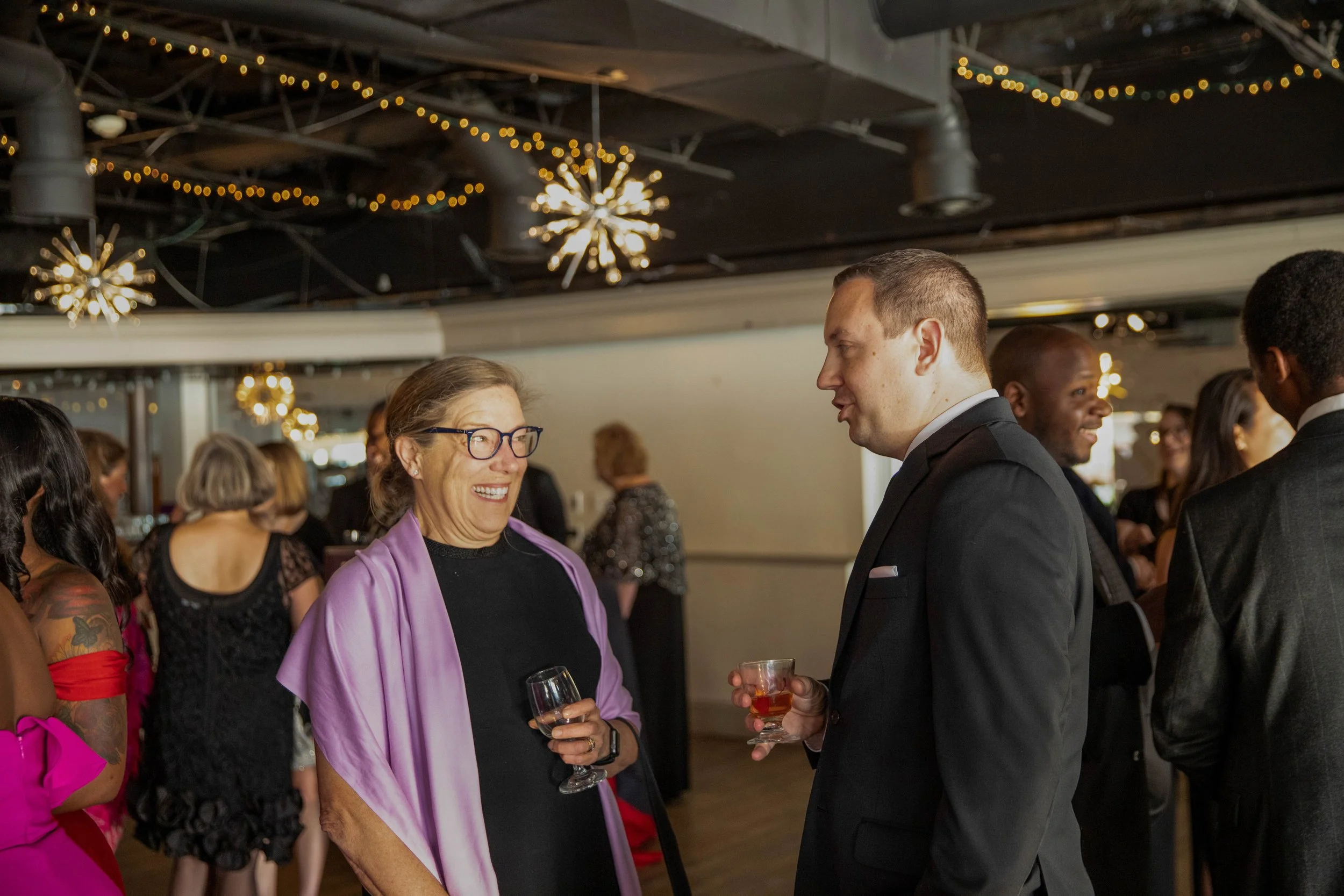 Two people in formal attire chatting at a social event, holding glasses of drinks, with warm lighting and other attendees in the background.