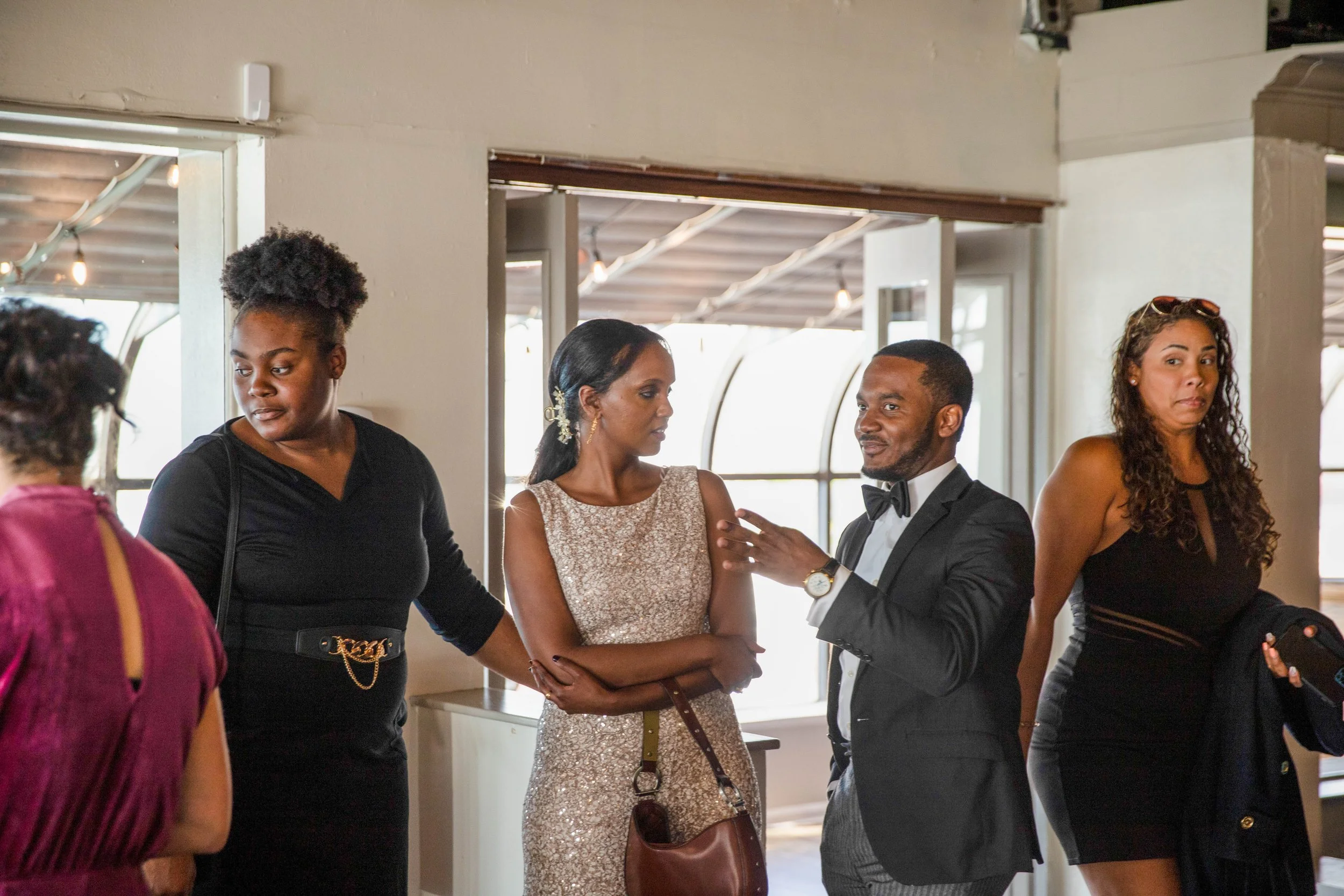 Group of people dressed in formal and semi-formal attire, standing indoors with large windows in the background, engaging in conversation.