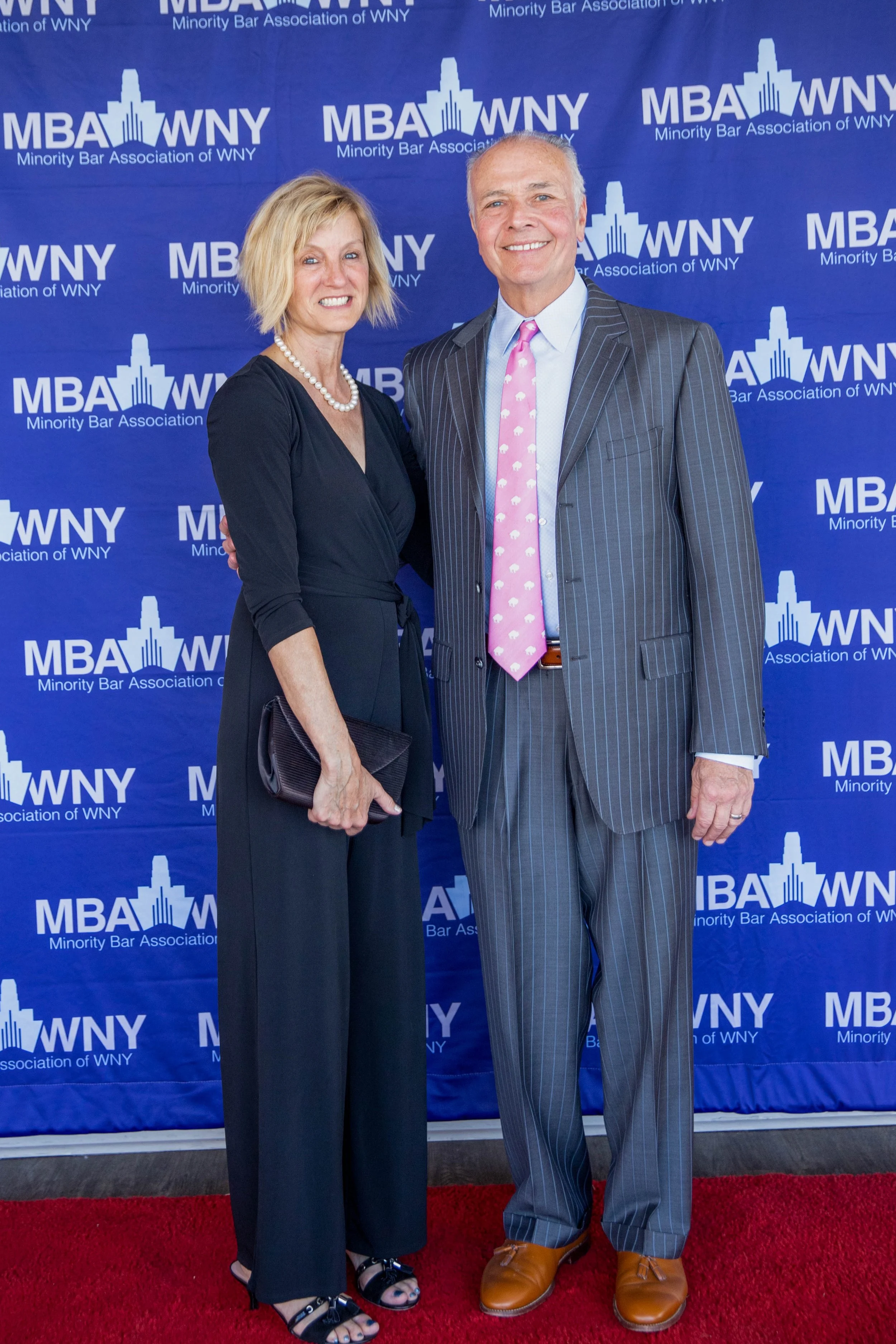 Two professionally dressed individuals, a woman and a man, standing on a red carpet in front of a blue backdrop with the logo and text for the Minority Bar Association of WNY. The woman is wearing a black dress with a pearl necklace, holding a black 