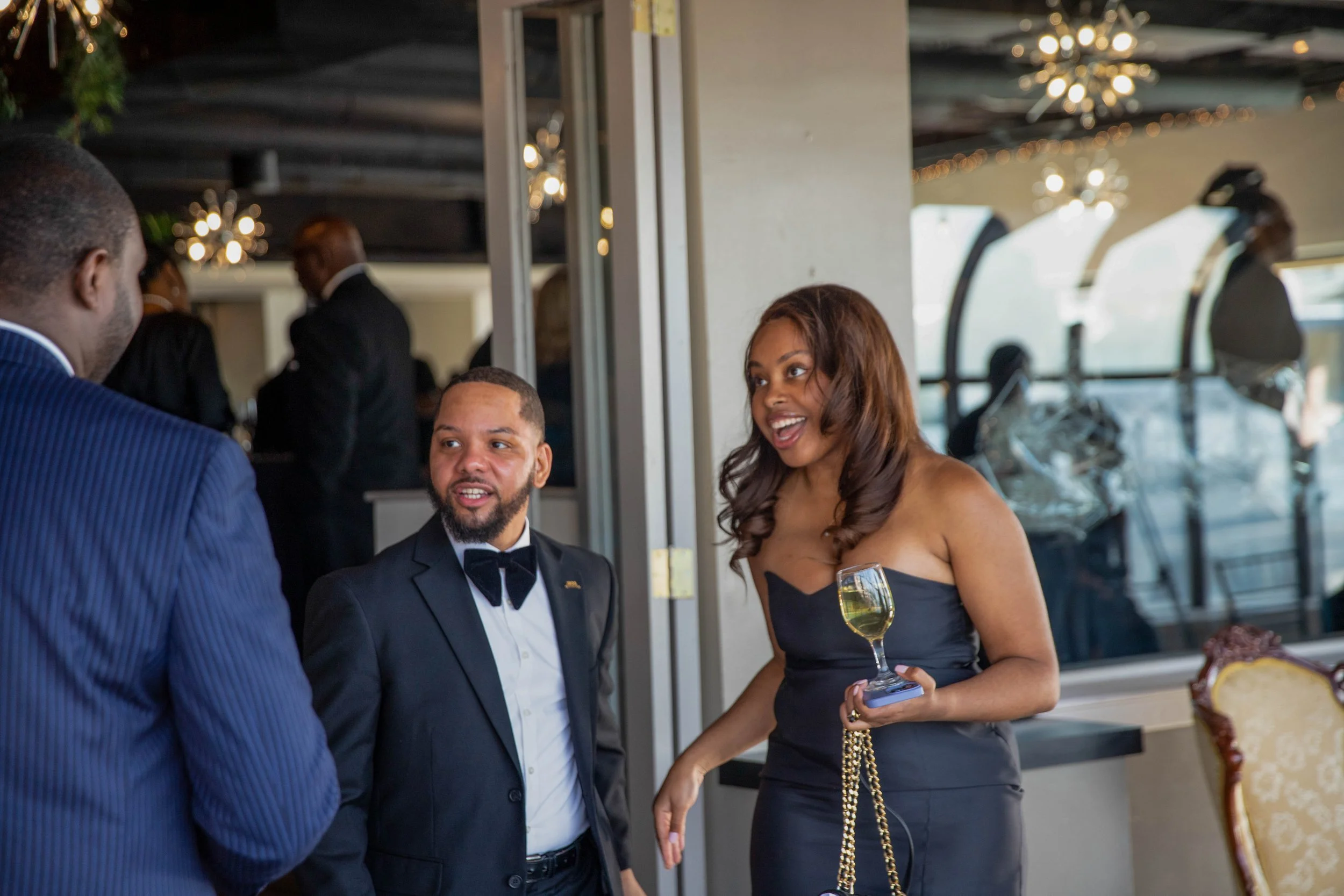 A diverse group of three people, two men and one woman, dressed formally at an event, engaged in conversation. The woman is holding a glass of champagne and a small tray, smiling, while the men are in suits.