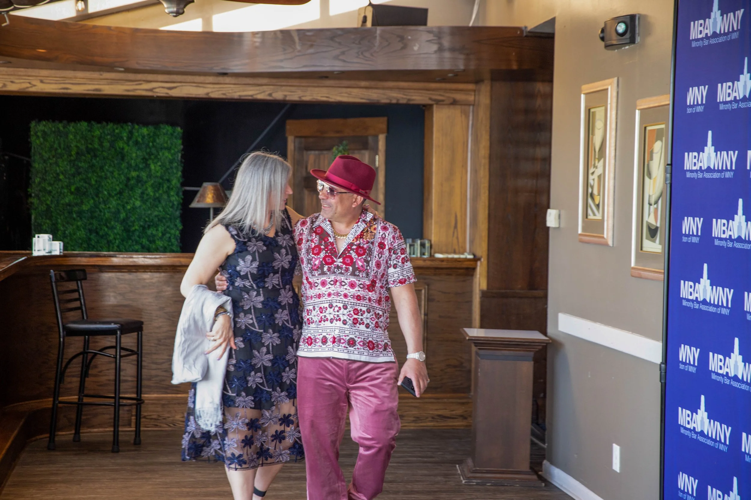 Two women walking together in an indoor setting, wearing colorful clothing, with one in a floral dress and the other in a patterned shirt and pink pants. There is a blue sign with white text on the right and a green wall decoration in the background.