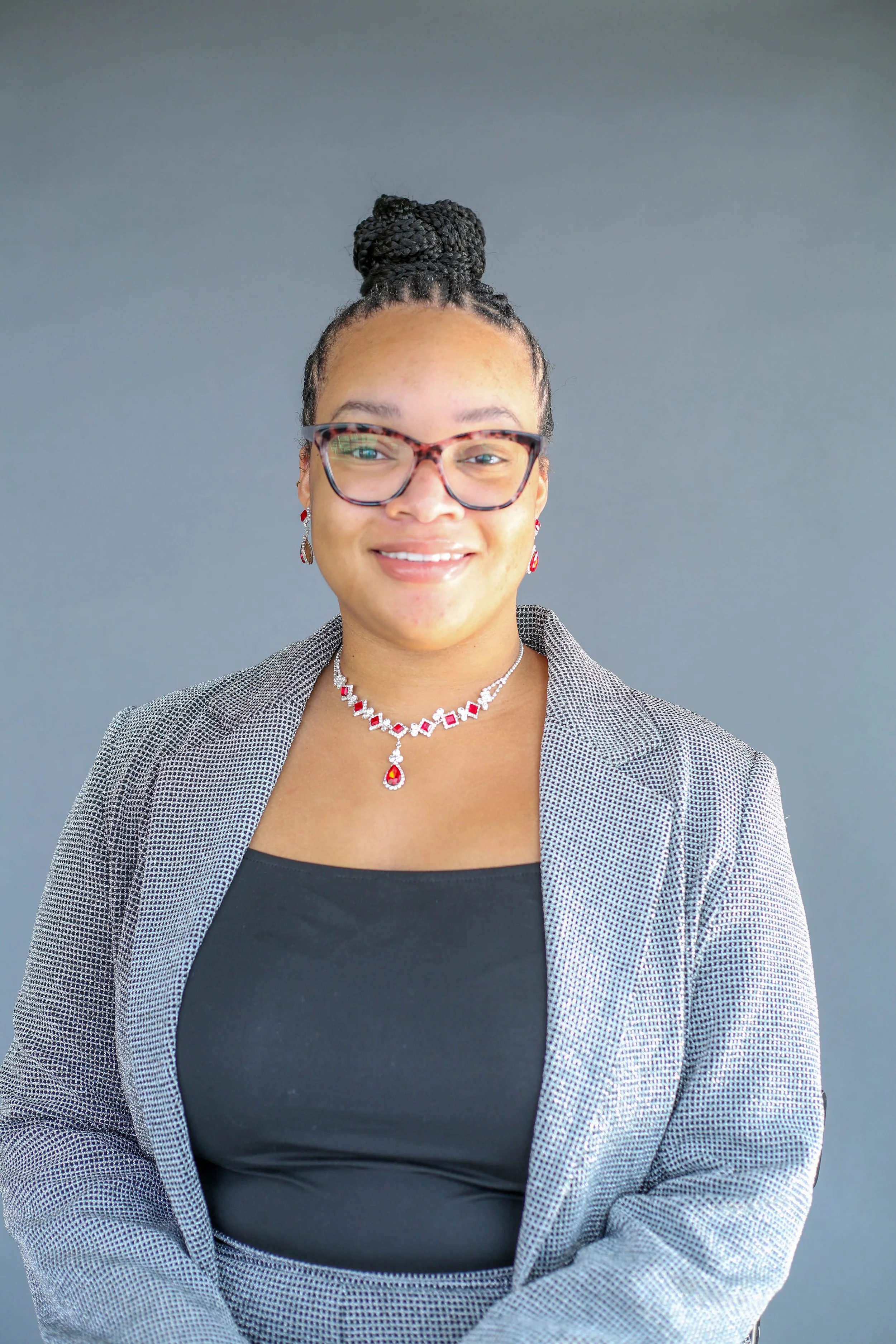 A woman with dark curly hair styled in a high bun, wearing glasses, a black top, and a checkered blazer, accessorized with red and silver jewelry, smiling in front of a plain gray background.
