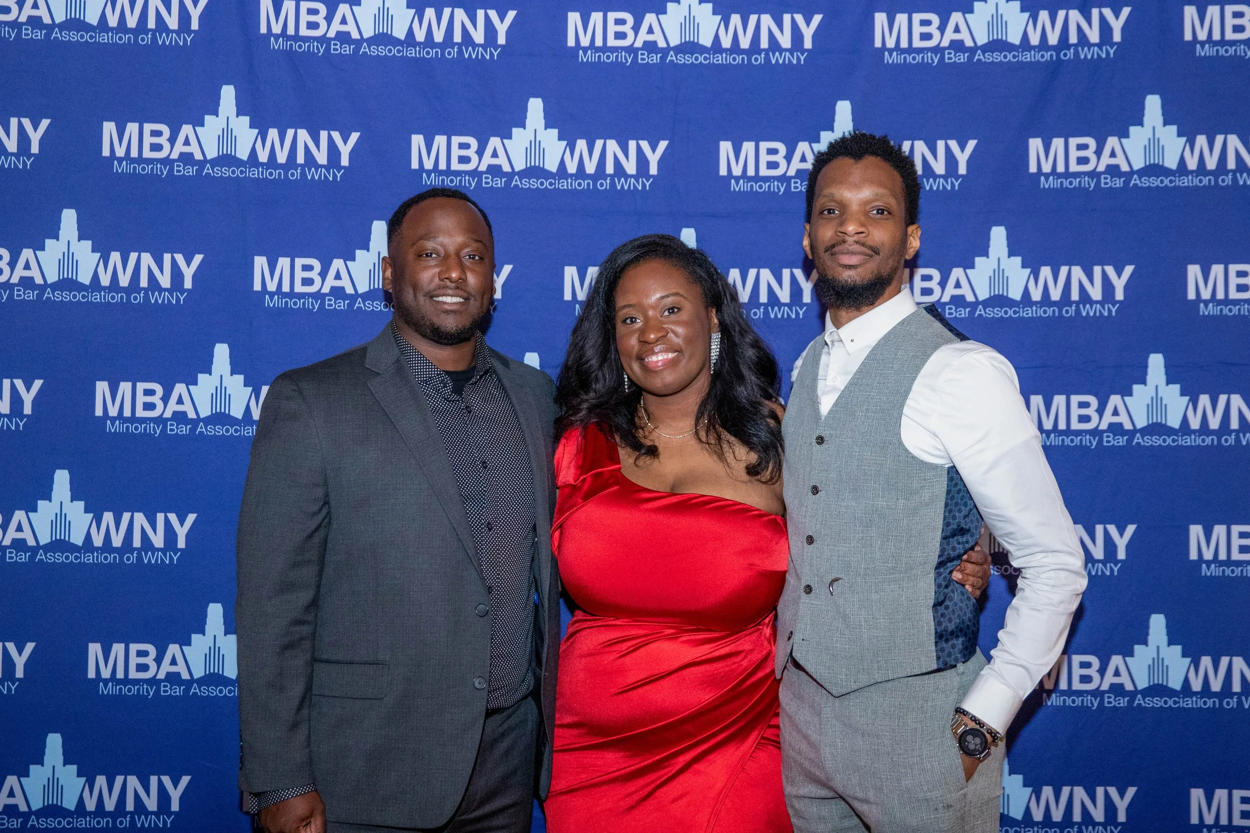 Three people standing in front of a blue backdrop with the logo and text of the Minority Bar Association of WNY, smiling at the camera. The man on the left is wearing a dark gray suit and polka dot shirt, the woman in the middle is wearing a red sati