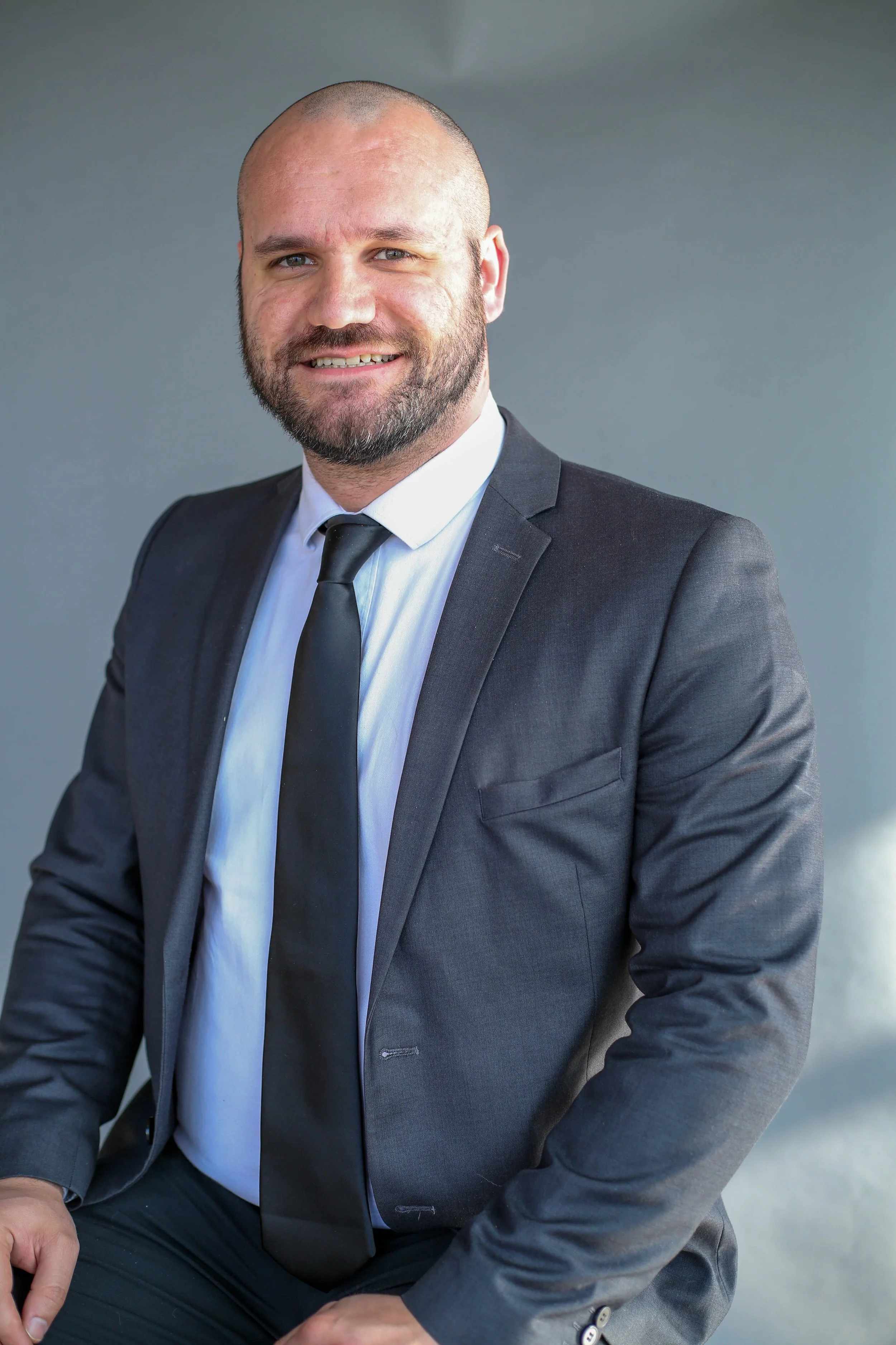 A man with a beard and short hair, dressed in a dark suit, white shirt, and black tie, sitting against a plain gray background.