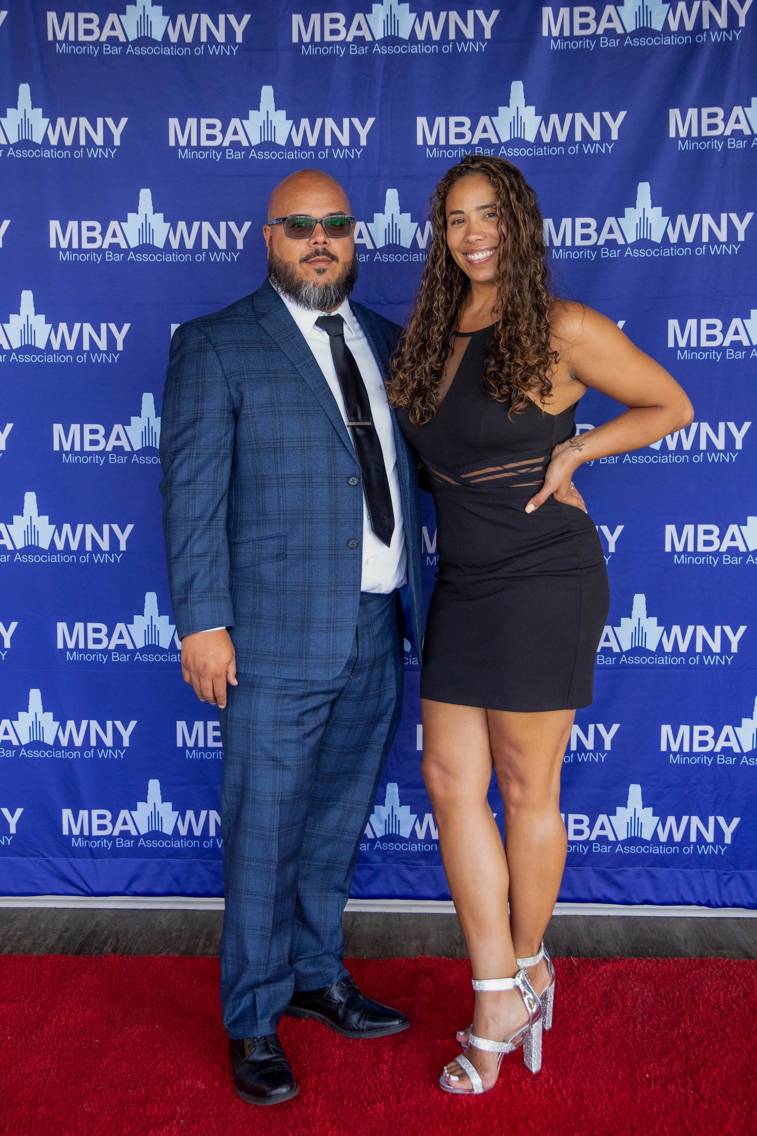 Two people standing on a red carpet at a formal event with a blue backdrop. The backdrop has the logo and text for the Minority Bar Association of WNY. The man on the left is wearing a blue plaid suit, white shirt, black tie, glasses, and has a shave