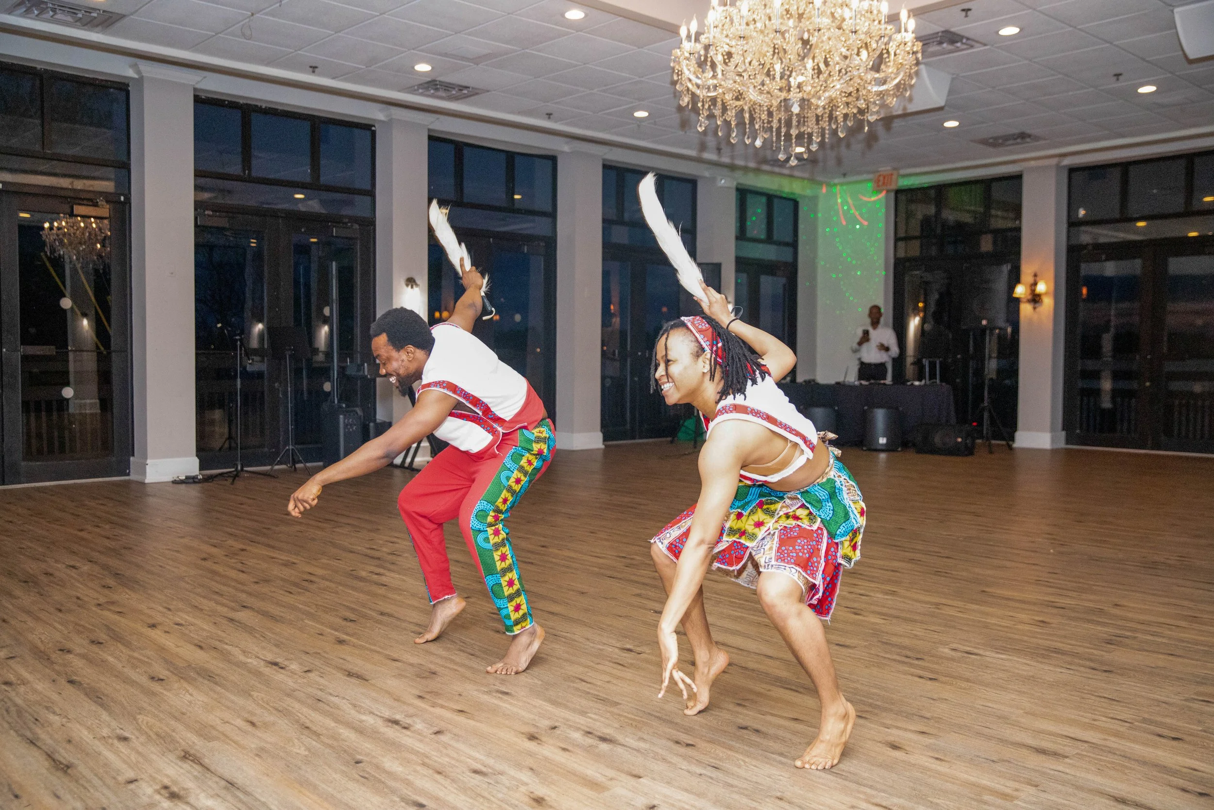 Two people dancing in traditional African attire, barefoot, in a spacious room with wooden floors, large windows, and a grand chandelier.