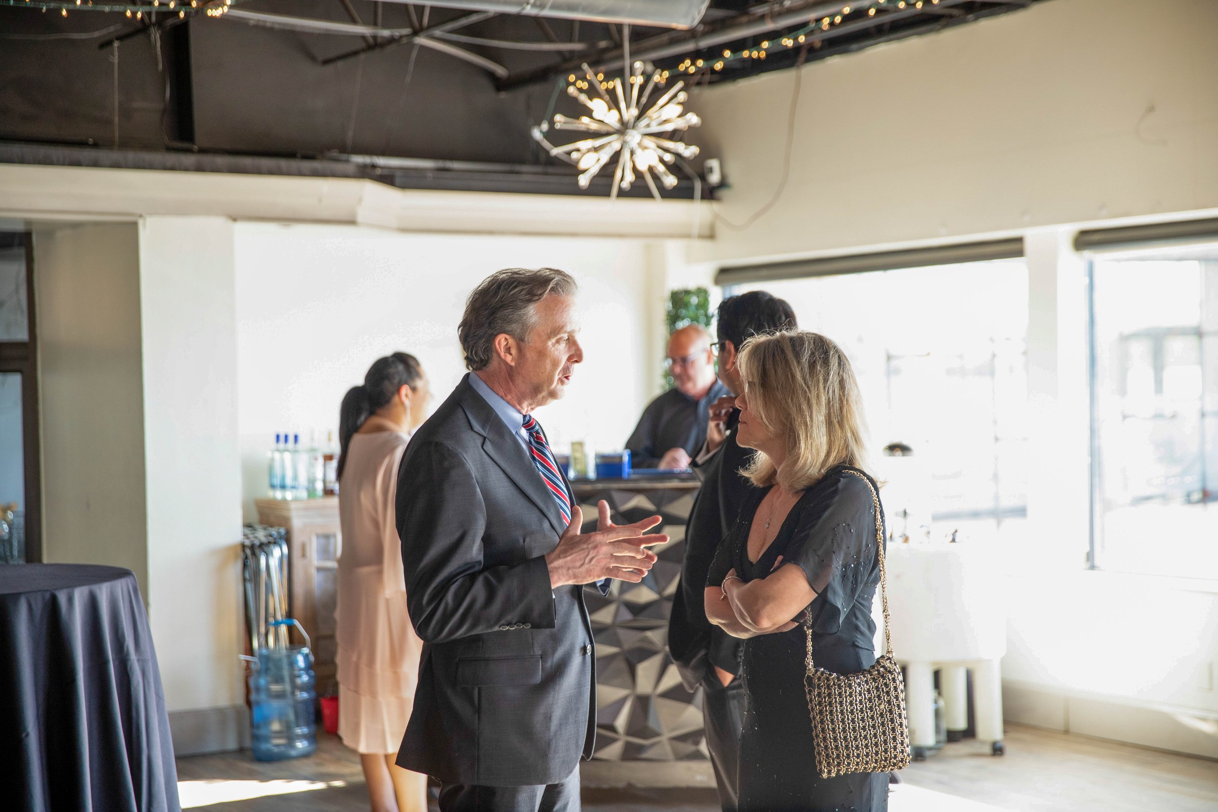 Two people, a man in a suit and a woman in a black dress, are engaged in conversation in a well-lit indoor space, with a woman in a pink dress and glasses in the background, along with others, and decorative lighting hanging from the ceiling.