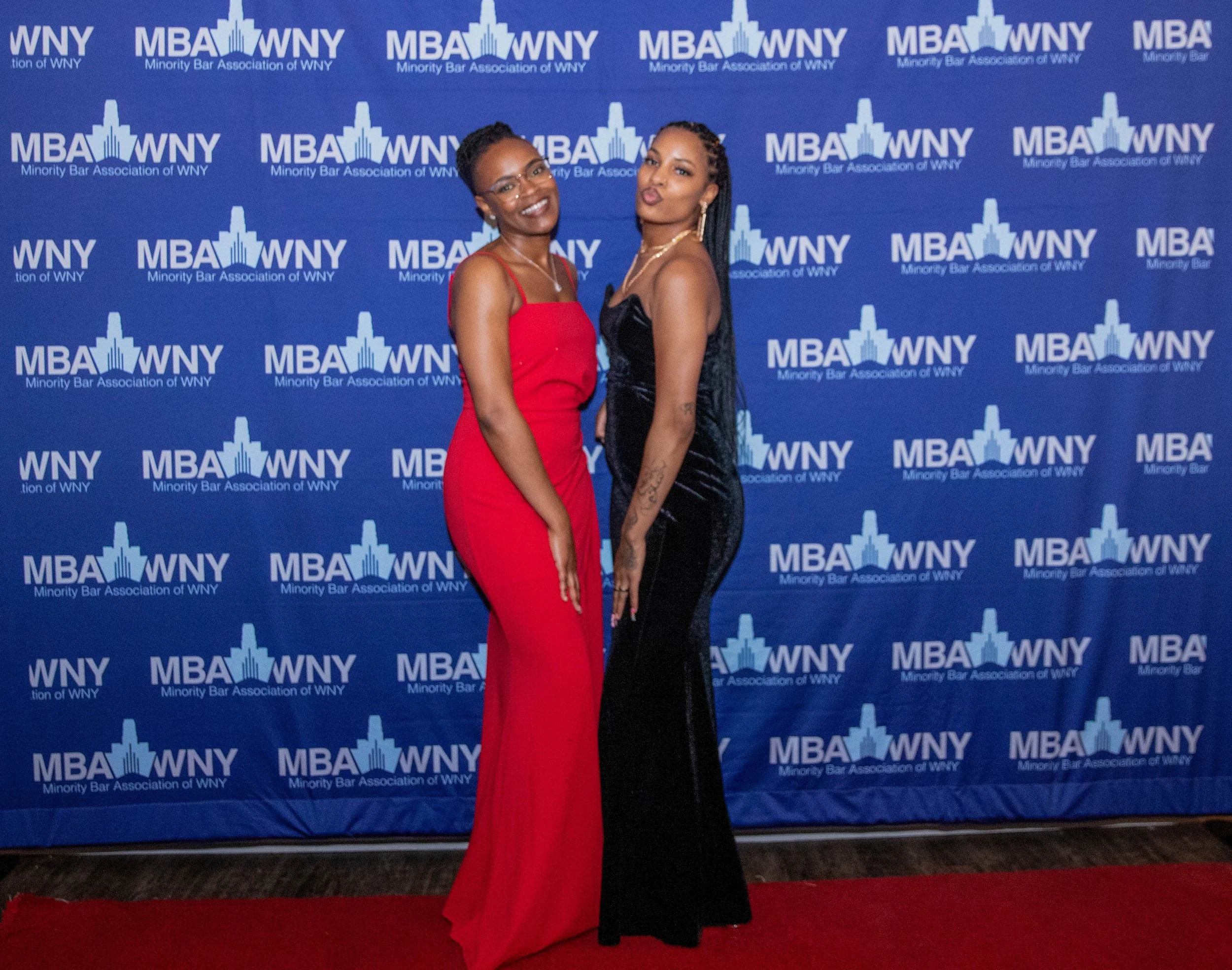 Two women dressed in evening gowns are standing in front of a blue backdrop with the logo and text for the Minority Bar Association of WNY. One woman is wearing a red dress and glasses, and the other is wearing a black dress with tattoos on her arm, 