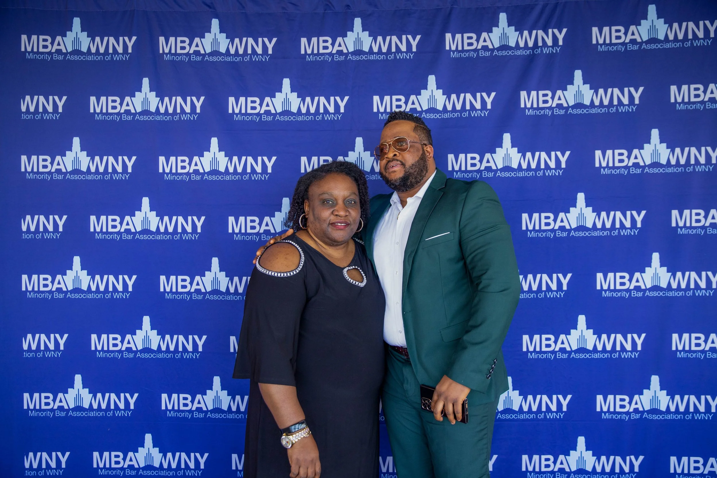 A woman and a man are posing together in front of a blue backdrop with white text and logos reading 'MBA WNY Minority Bar Association of WNY.' The woman is wearing a black dress with embellishments on the shoulders, and the man is wearing a green sui