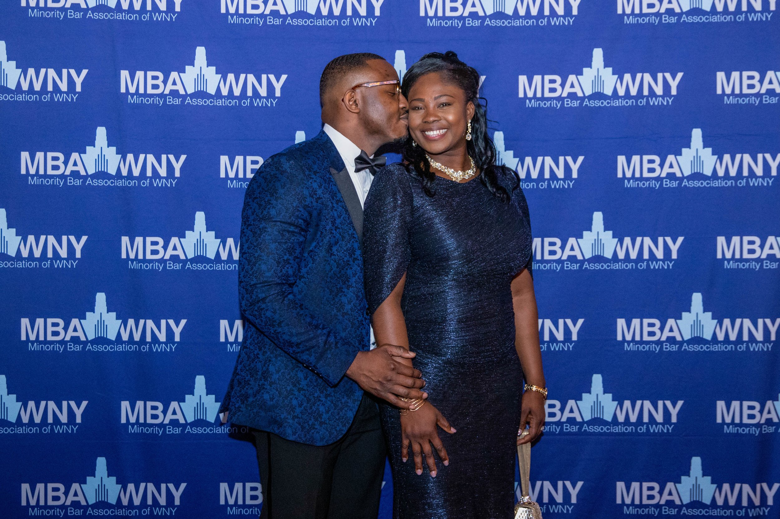 A man and woman at a formal event, with the man kissing the woman's cheek. They are dressed in formal attire and standing in front of a blue backdrop with white text and logos for the Minority Bar Association of WNY.