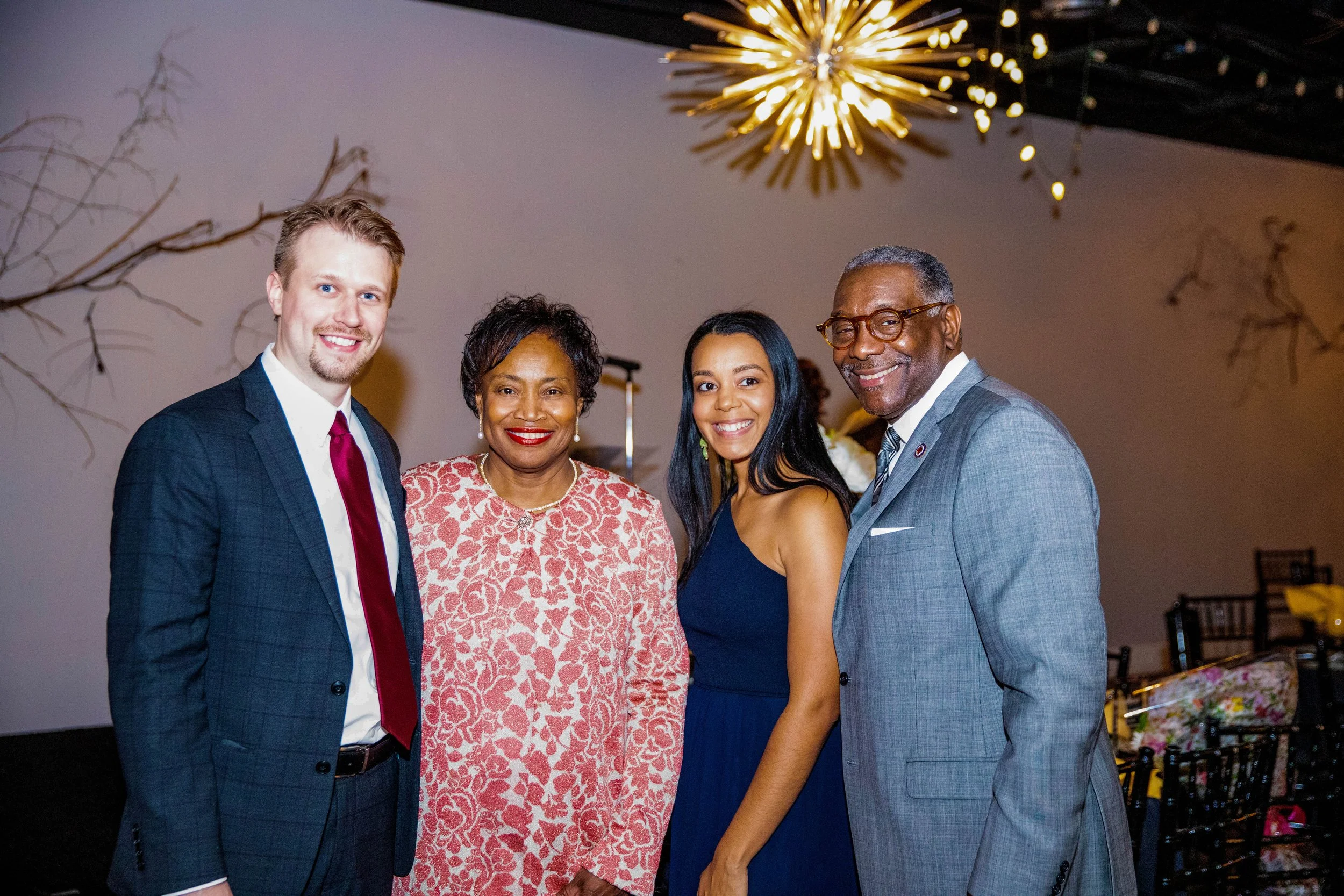 Group of four diverse people smiling at a social event, standing together indoors with artwork and decorative lights in the background.
