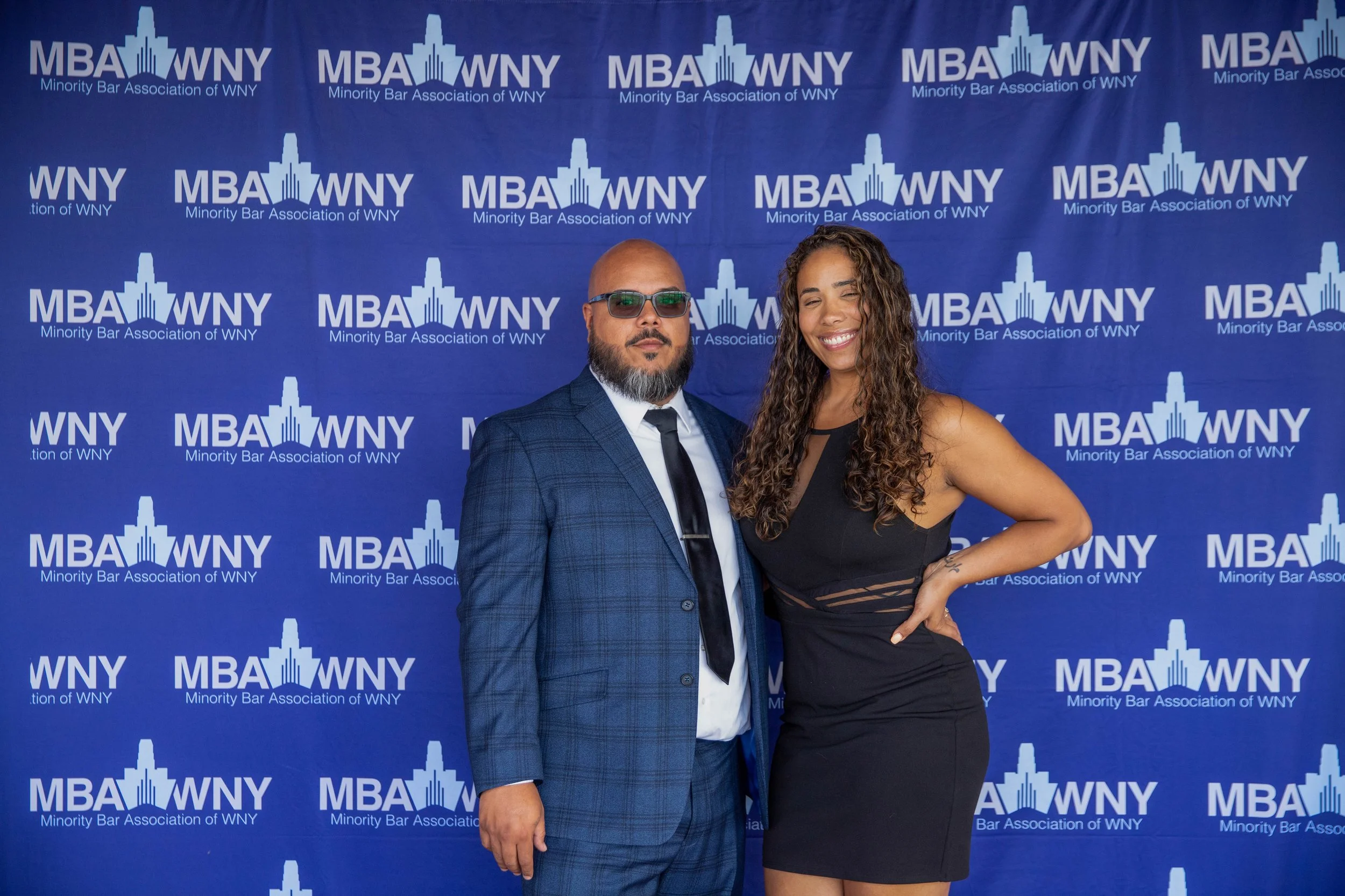 Two people standing in front of a blue backdrop with white text and logo that reads 'MBA WNY Minority Bar Association of WNY'. The man on the left is wearing a blue checkered suit, white shirt, black tie, sunglasses, and has a beard. The woman on the