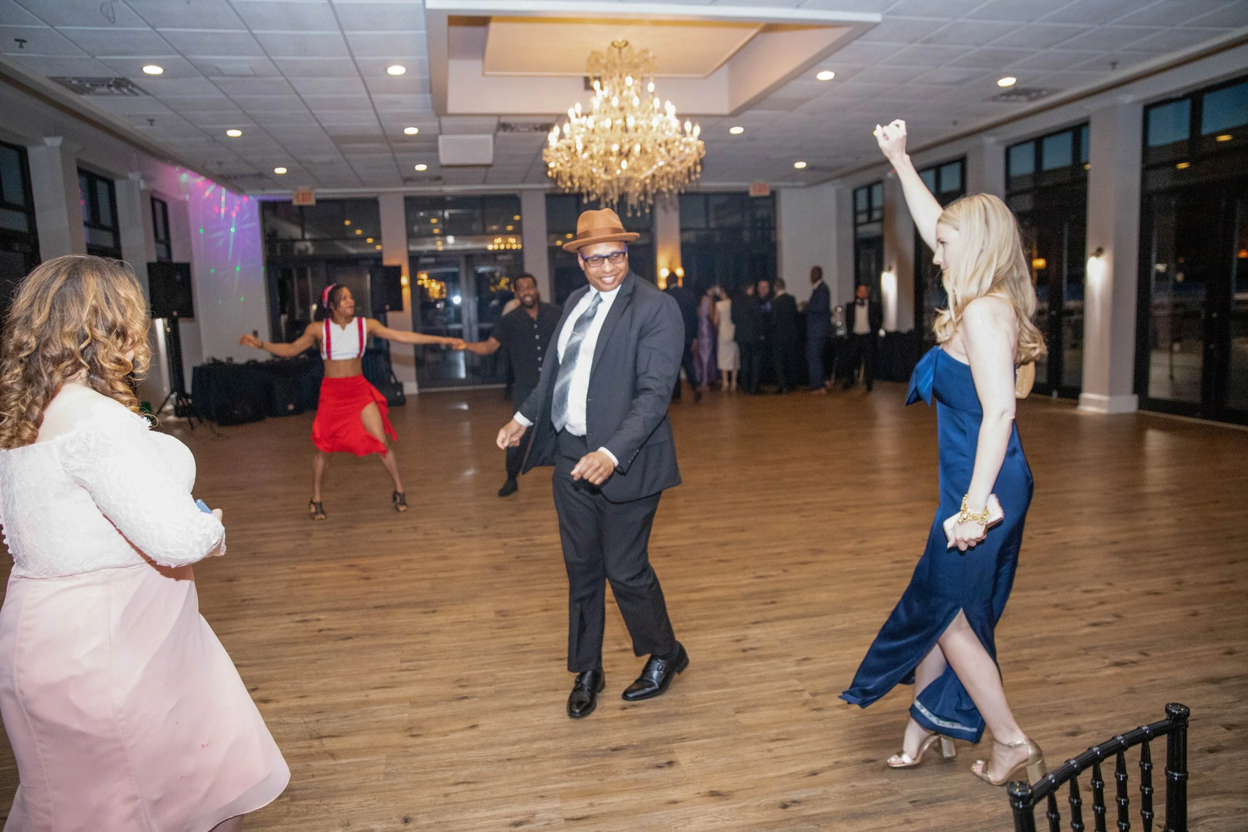 People dancing at an indoor celebration with a chandelier, windows, and wooden flooring.