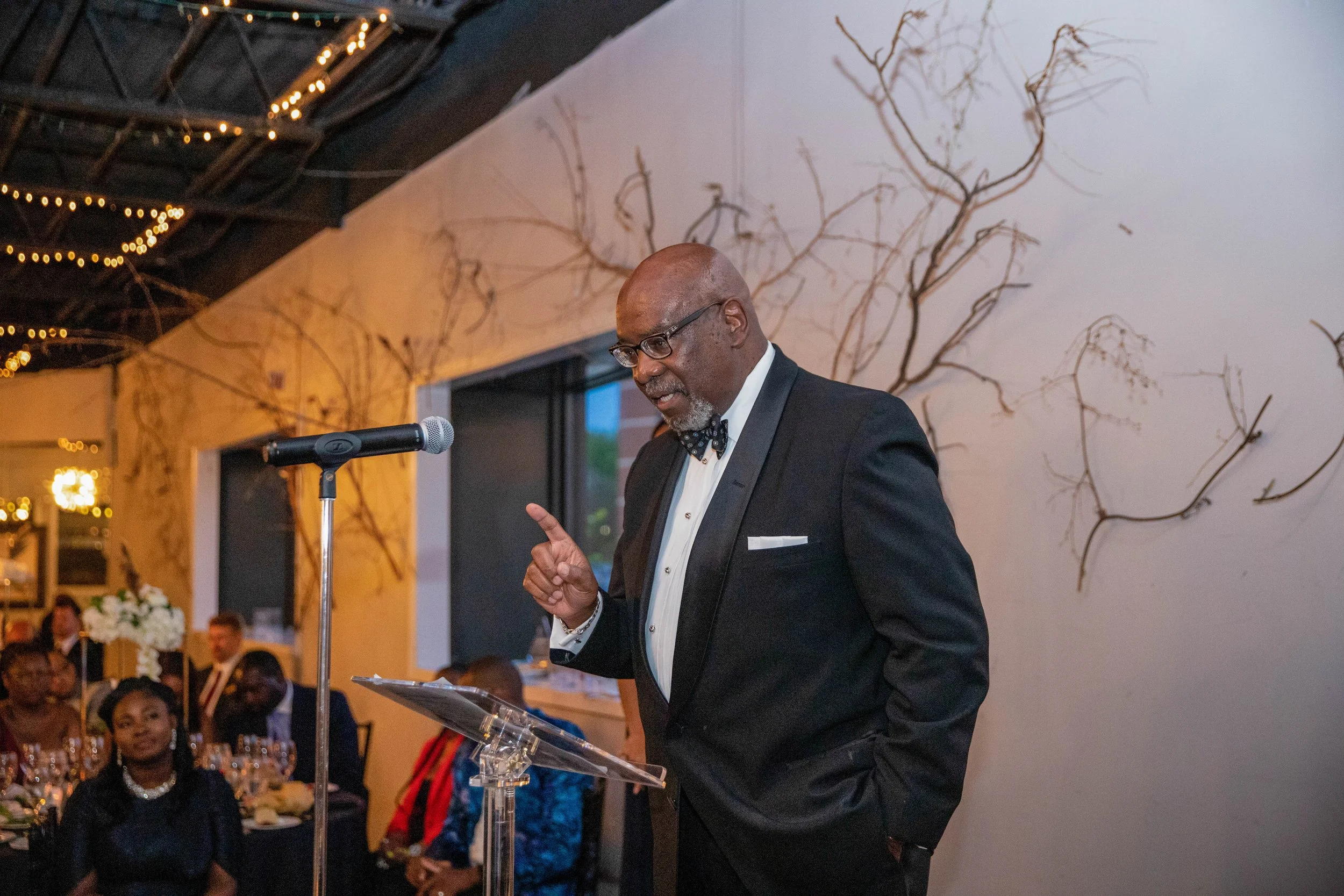 An elderly man in a tuxedo with a bow tie and glasses is speaking at a podium during a formal event. The background shows decorated walls with bare branches and string lights, and seated guests are visible in the background.