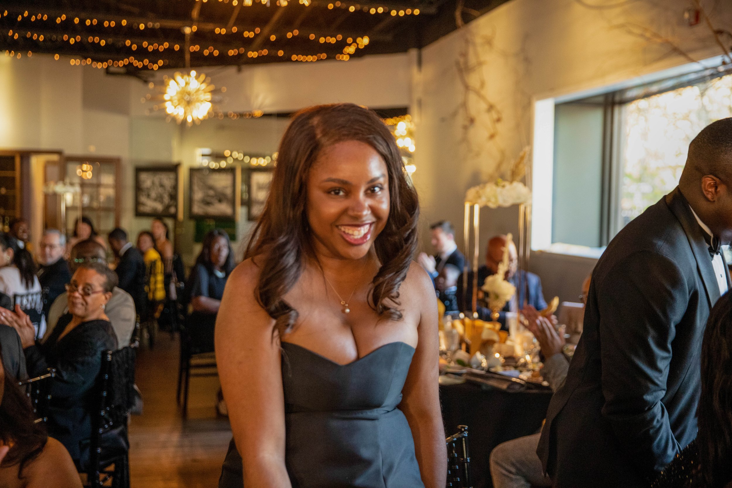 A smiling woman with brown wavy hair wearing a strapless black dress at a festive gathering with holiday decorations and guests in the background.